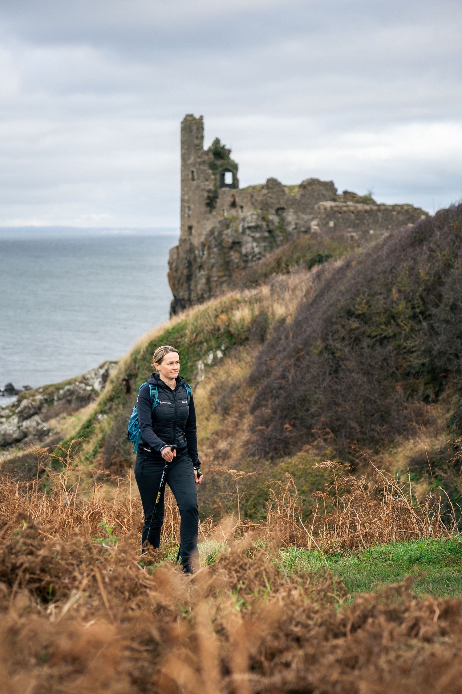 Woman hiker with backpack walking coastal path near ancient castle ruins overlooking the sea on overcast day.
