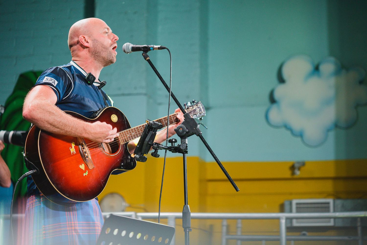 Bald man playing a sunburst acoustic guitar and singing into a microphone outdoors at a live music event.