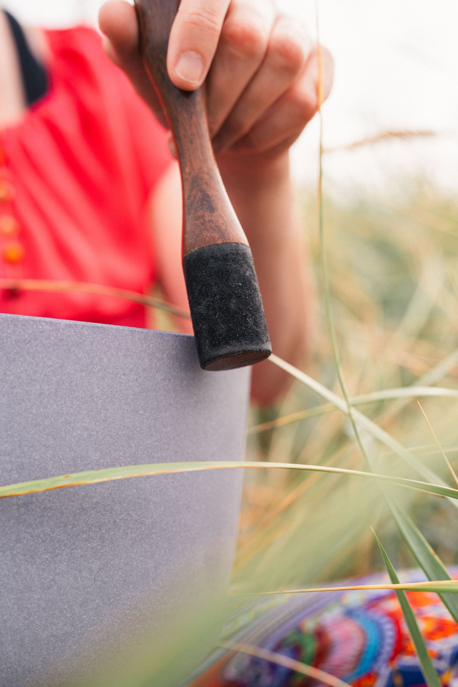 Close-up of a hand holding a wooden mallet against a gray singing bowl outdoors in tall grass.