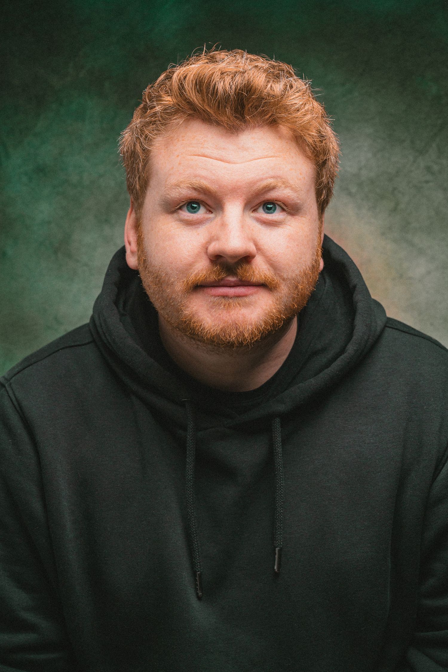 Red-haired bearded man in a black hoodie posing for a portrait against a dark green textured background.