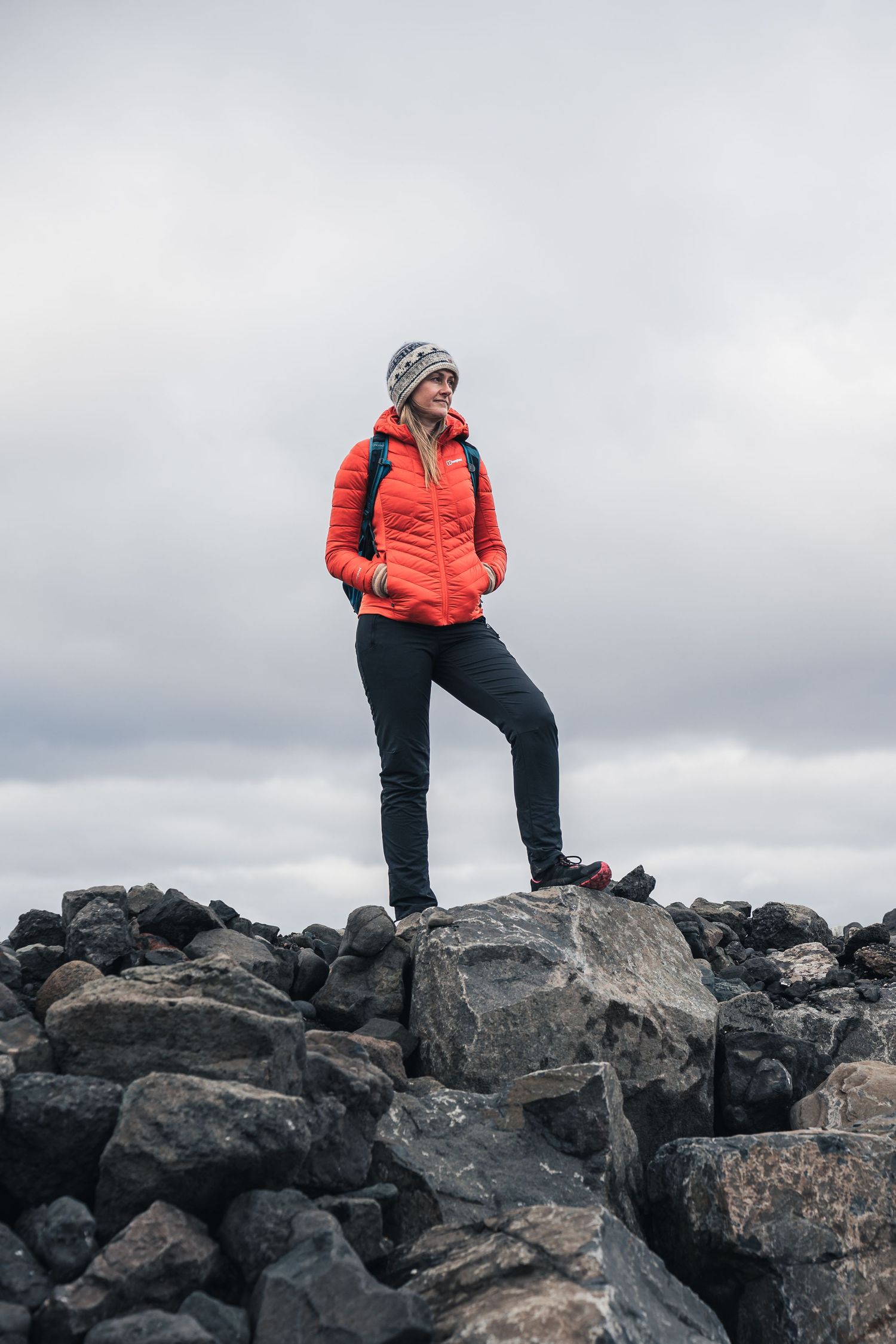 Woman in red jacket and beanie standing on rocky terrain against cloudy sky, hiking outdoors.