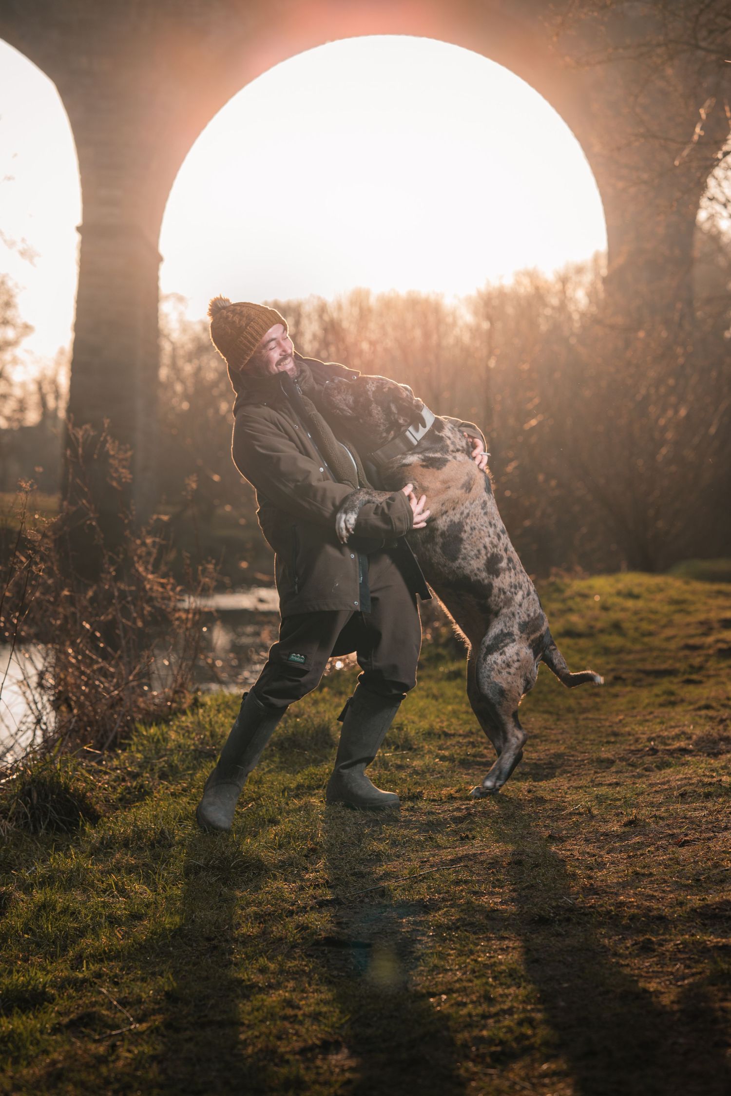 Man playing with a large spotted dog outdoors under a stone arch at sunset.