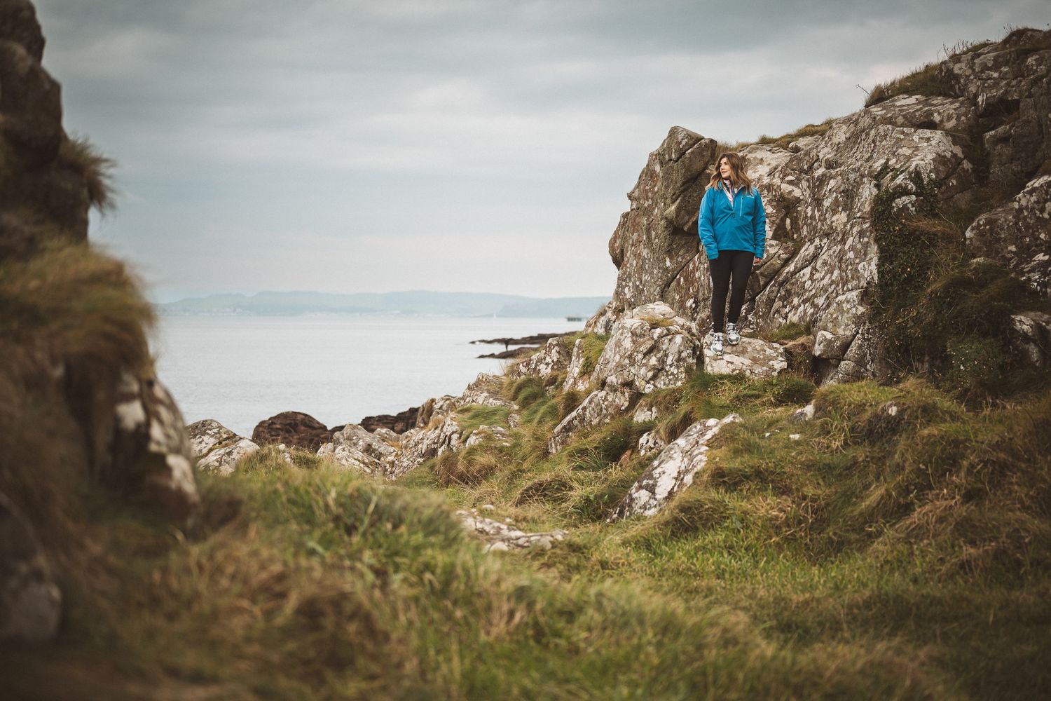 A hiker in a blue jacket stands among rocky terrain with a coastal ocean view in the background.