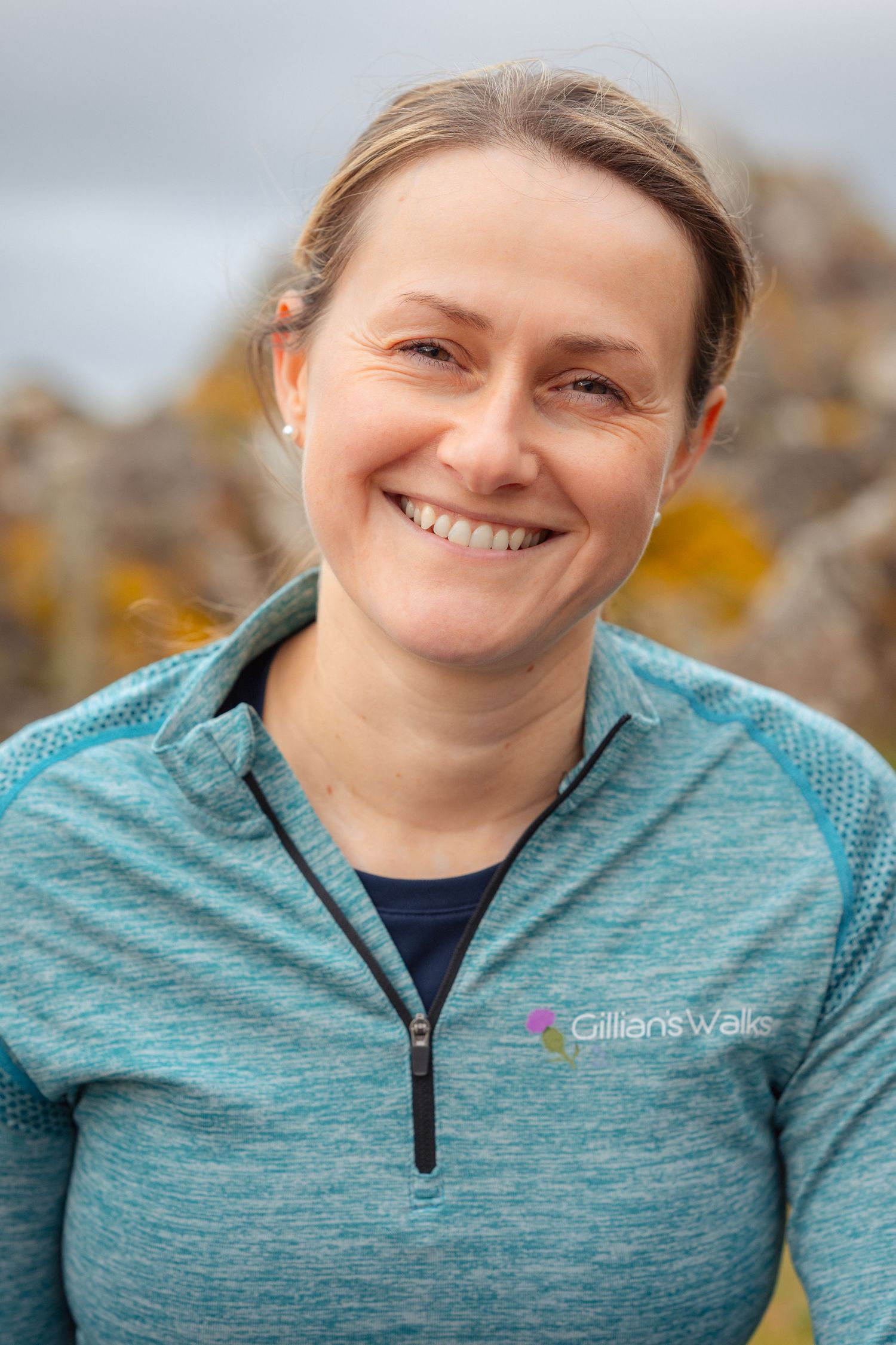 Smiling woman wearing a teal Gillians Walks zip-up top outdoors with rocky background.