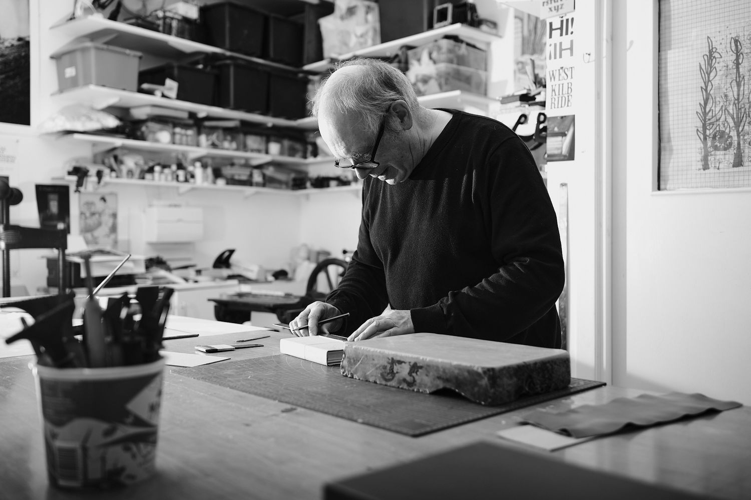Black and white photo of an elderly man working at a wooden desk in a cluttered art studio.