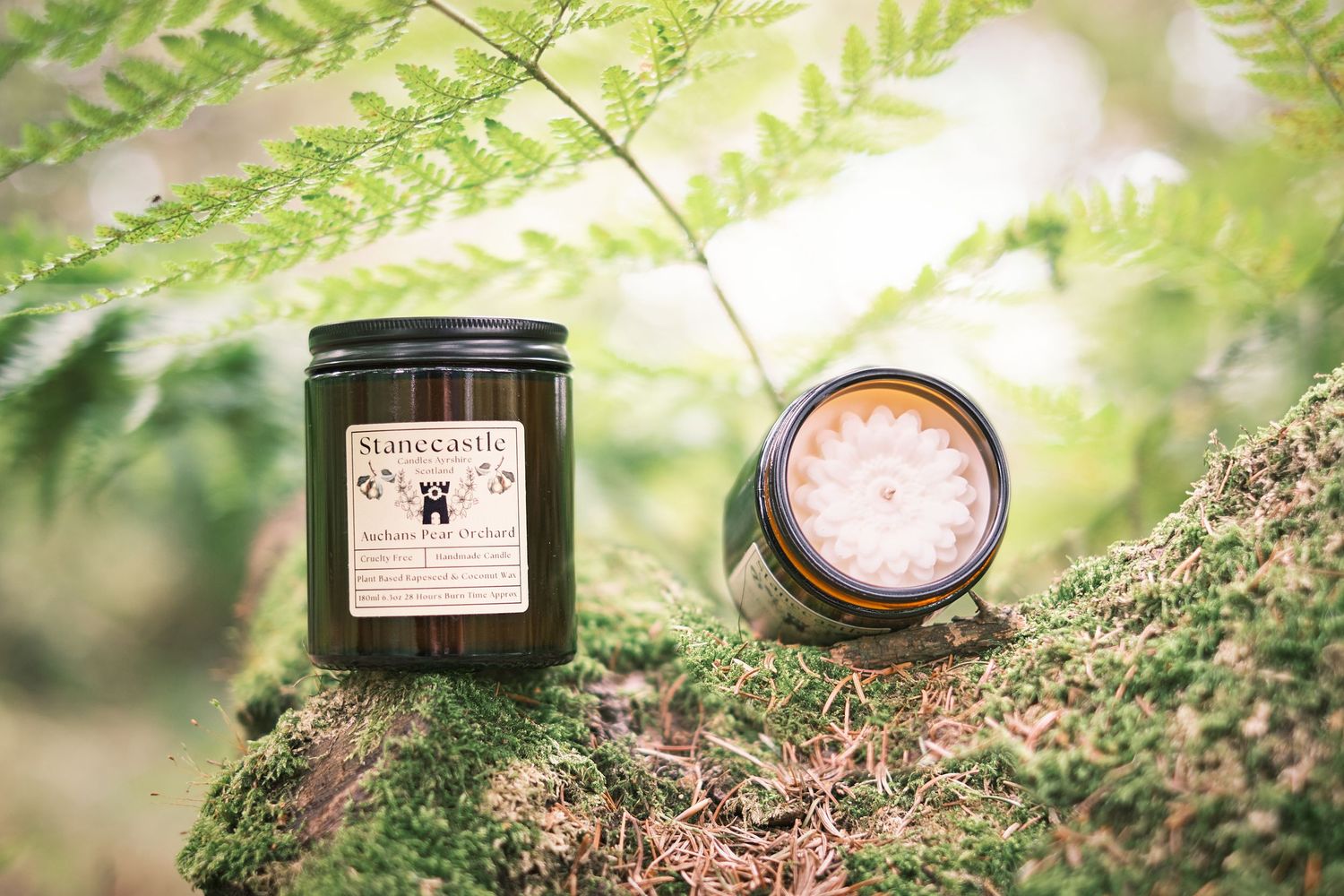 Two amber glass candle jars on a mossy rock surrounded by lush green ferns in a natural outdoor setting.