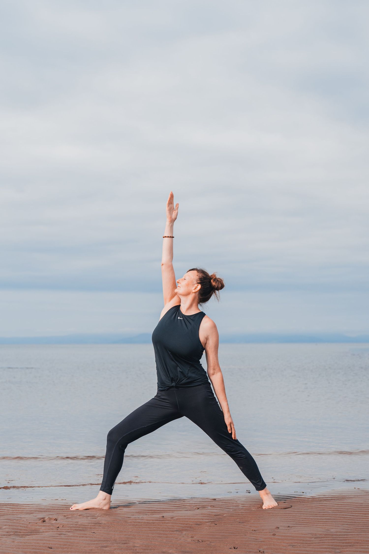 Woman in black athletic wear practicing warrior yoga pose on a sandy beach with calm ocean backdrop.