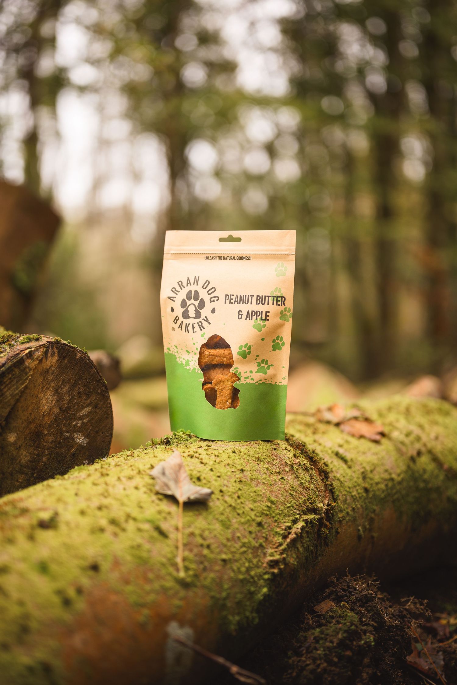 Arran Dog Bakery peanut butter and apple dog treats bag resting on a mossy log in a forest setting.