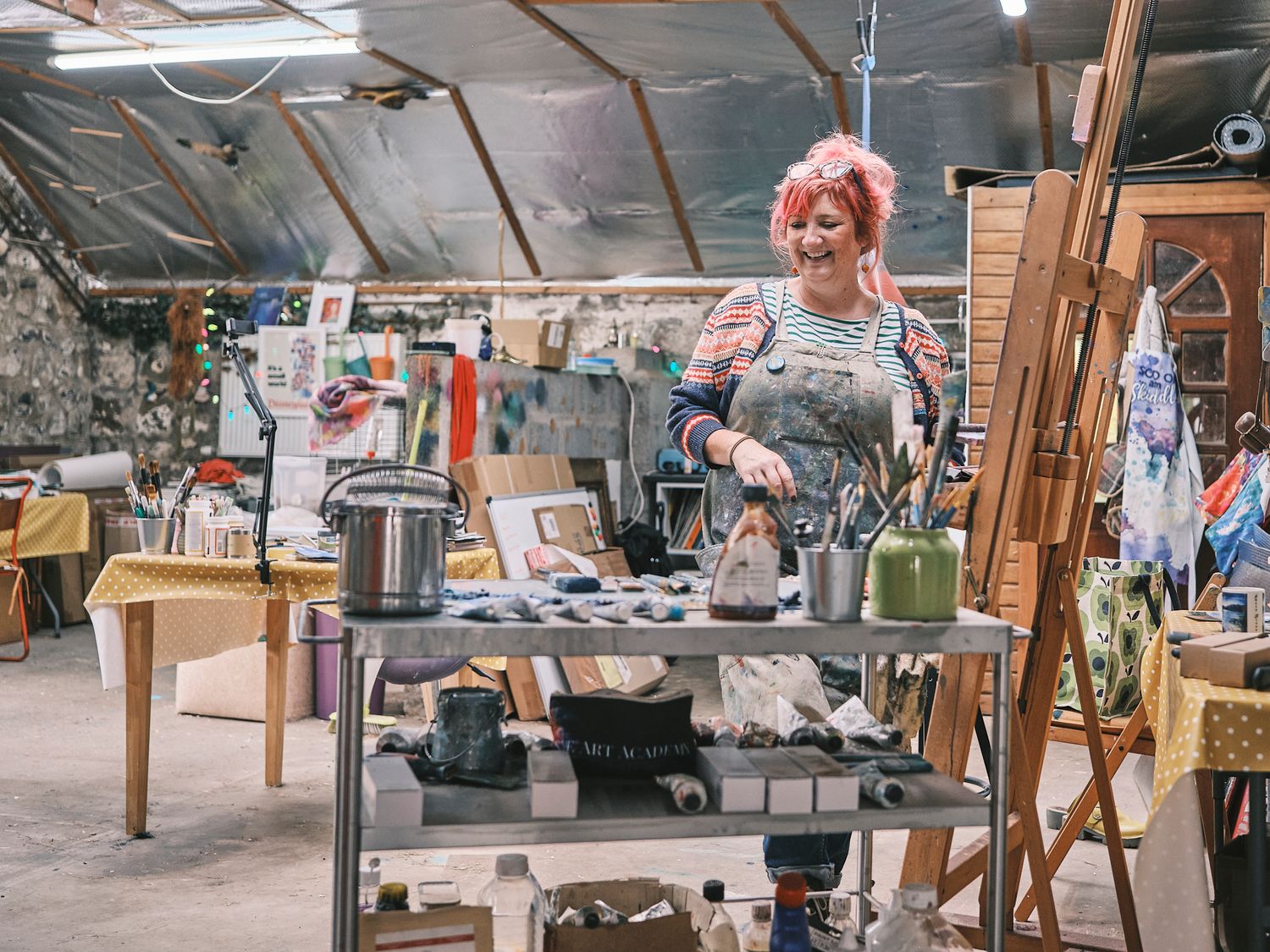 Artist with pink hair works at a cluttered studio table surrounded by art supplies and easels in a creative workspace.