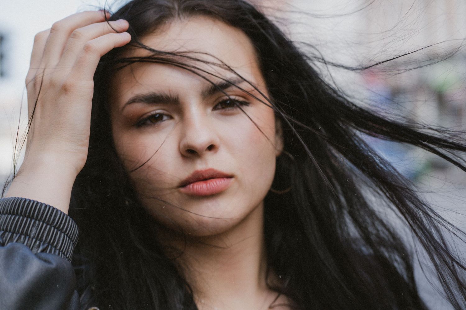 Young woman with long dark hair blowing in wind, hand raised to head, wearing leather jacket outdoors.