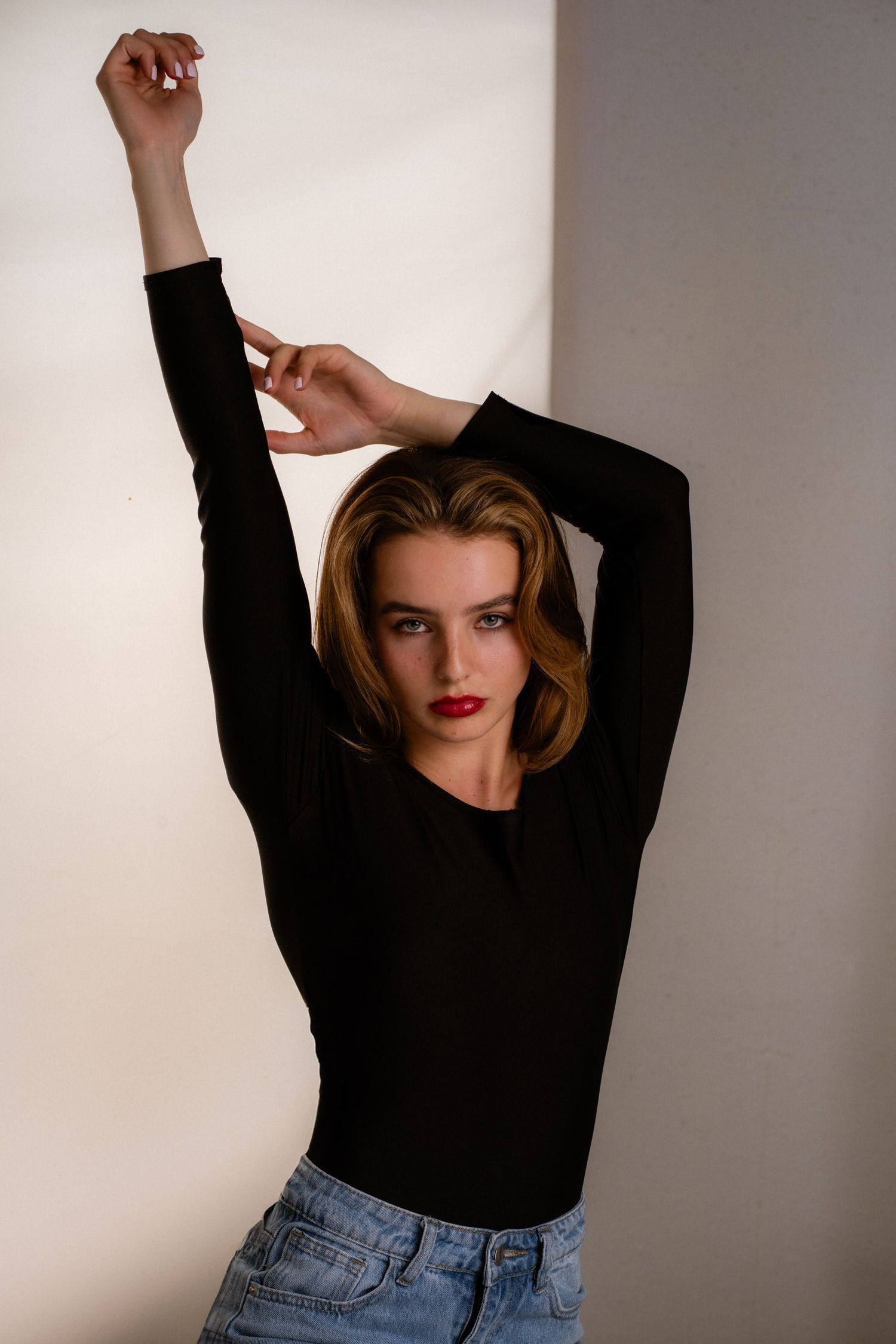 Young woman in black long-sleeve top and jeans poses with arms raised against a white wall.