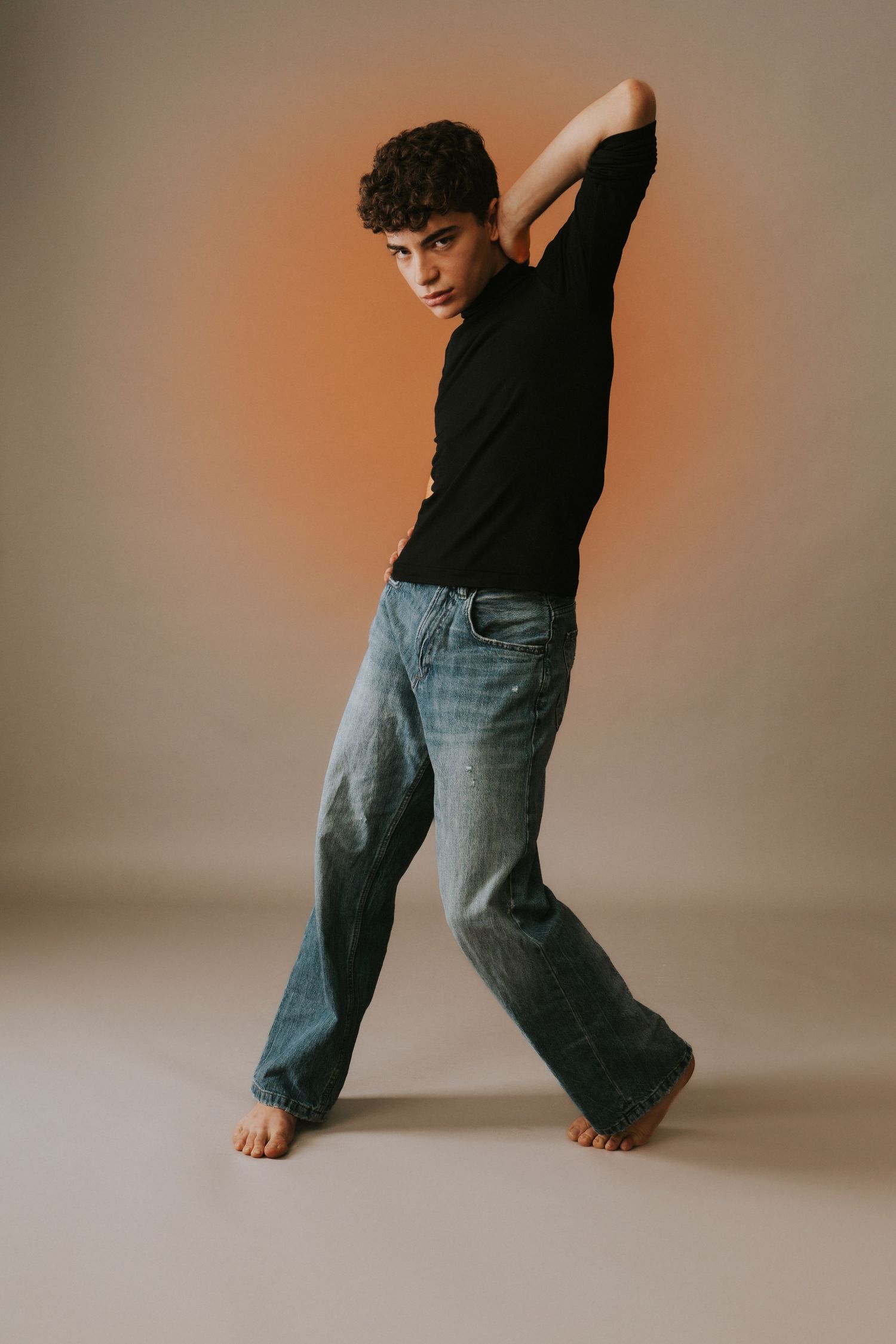 Young man in black t-shirt and wide-leg jeans posing barefoot on orange studio background.
