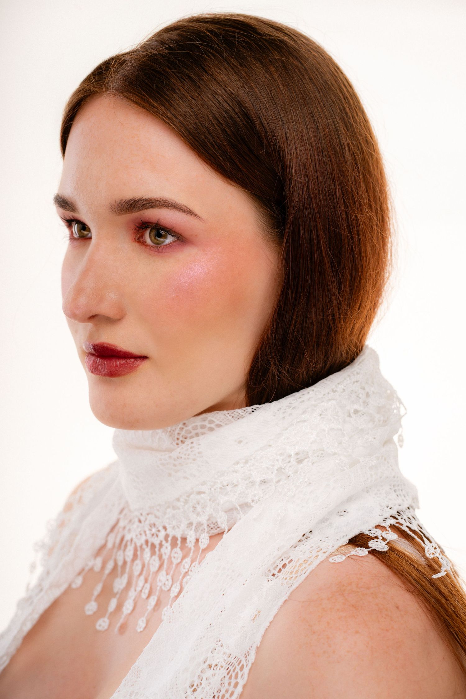 Young woman with auburn hair wearing white lace scarf and bold burgundy lip makeup in studio portrait.