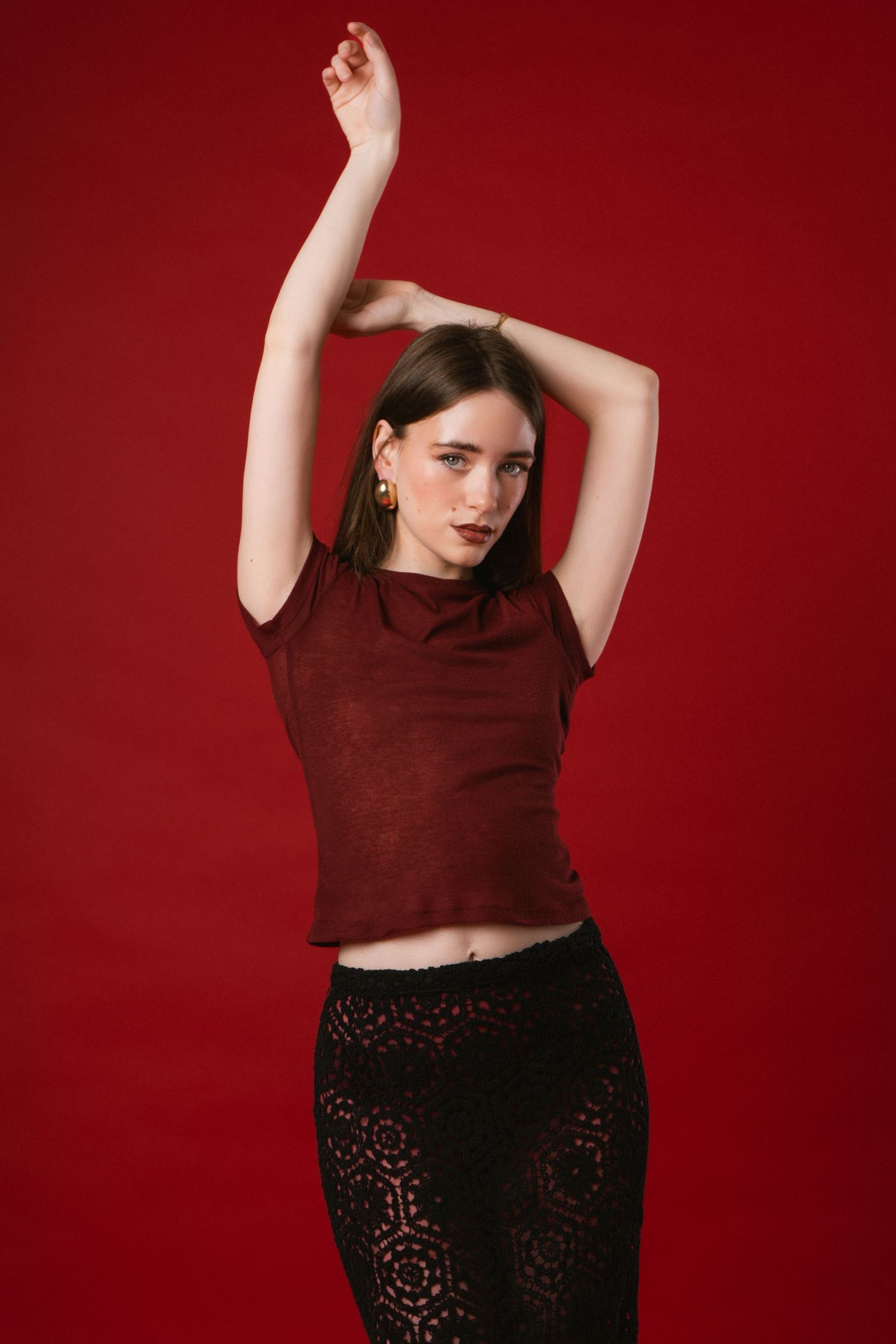 Young brunette woman in burgundy crop top and black lace skirt poses with arms raised on red background.