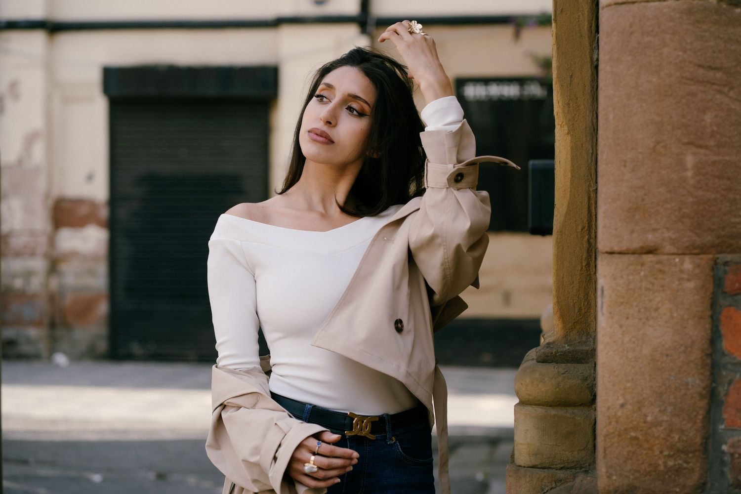 Young woman in white off-shoulder top and trench coat posing confidently against a stone column on a city street.