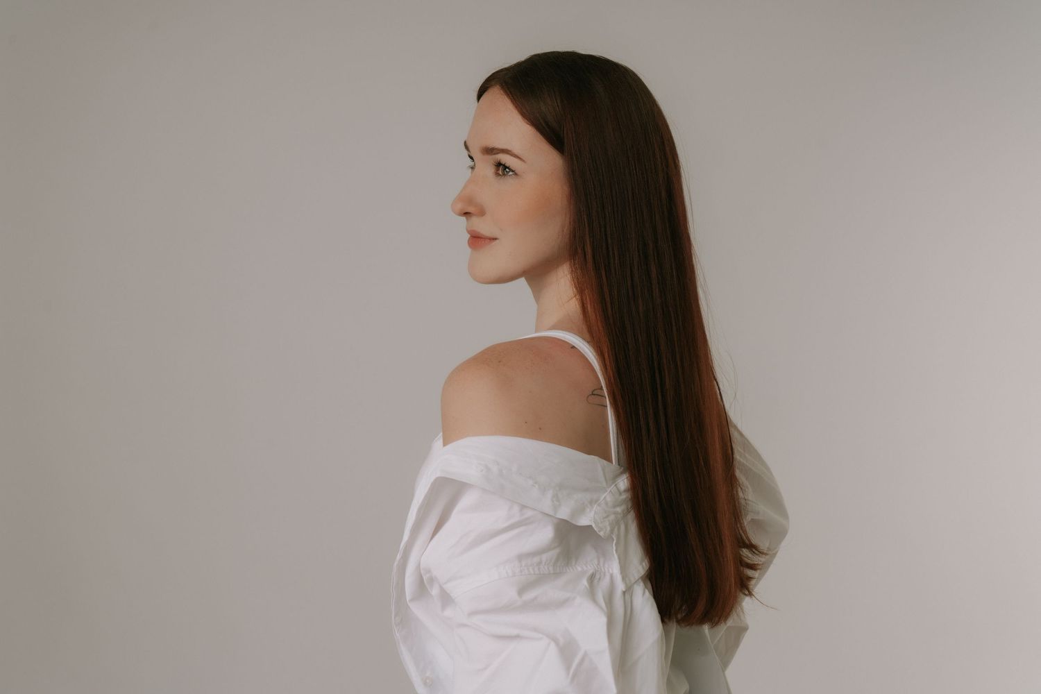 Young woman with long brown hair in white off-shoulder shirt, side profile view, light gray background.