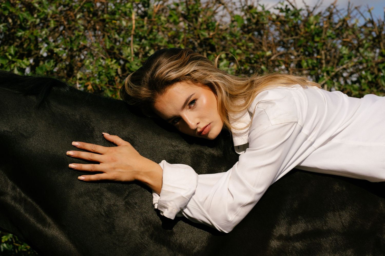 Woman in white blouse resting her head on a dark horse, surrounded by lush green foliage outdoors.