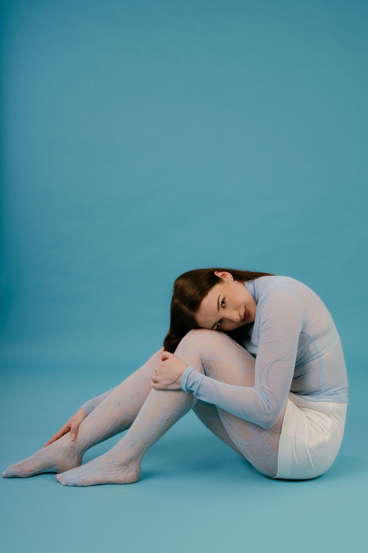 Young woman in blue mesh turtleneck hugging her knees while sitting on blue studio background.