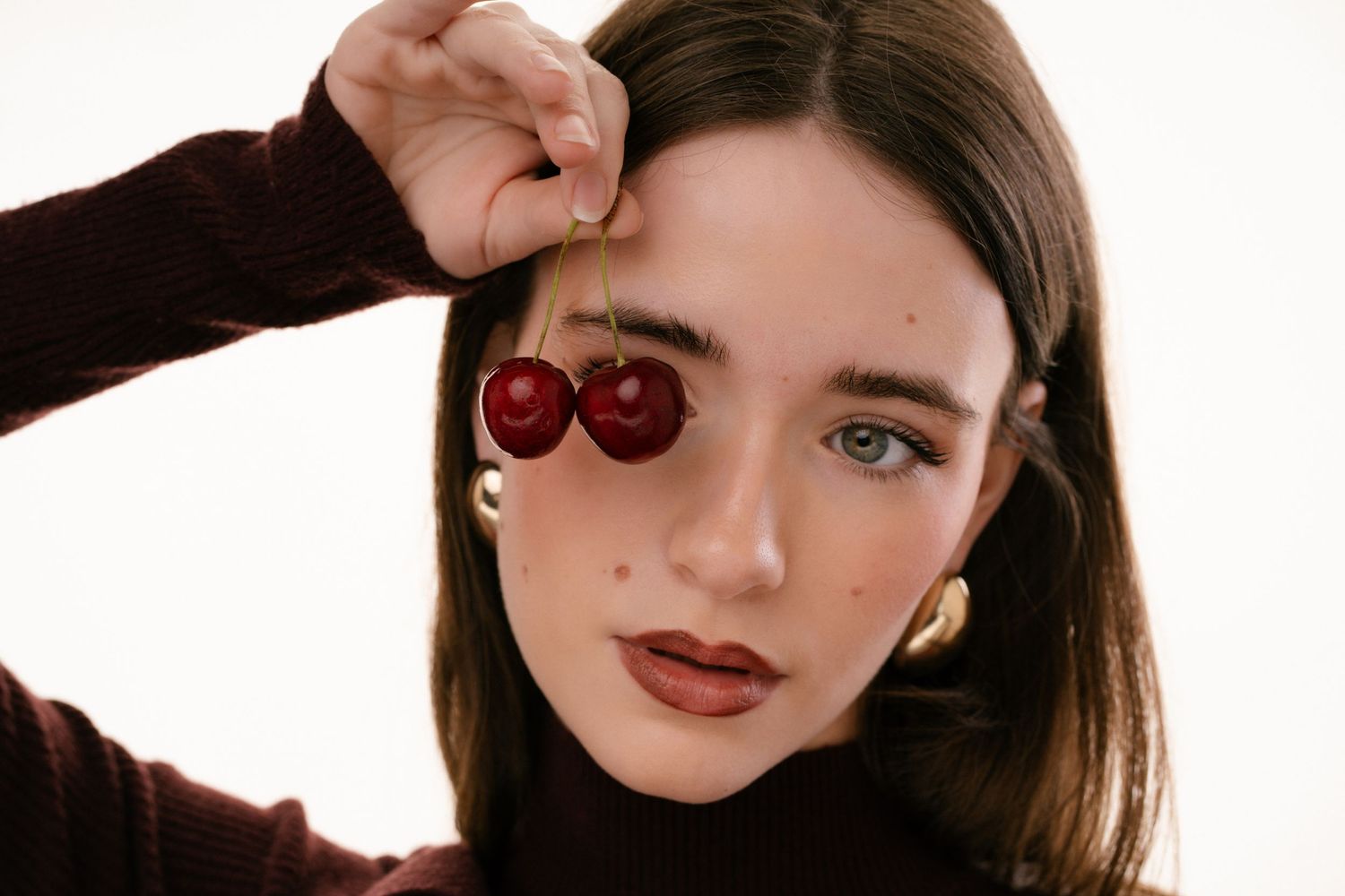 Close-up portrait of young woman holding two red cherries over her eye, wearing gold hoop earrings.