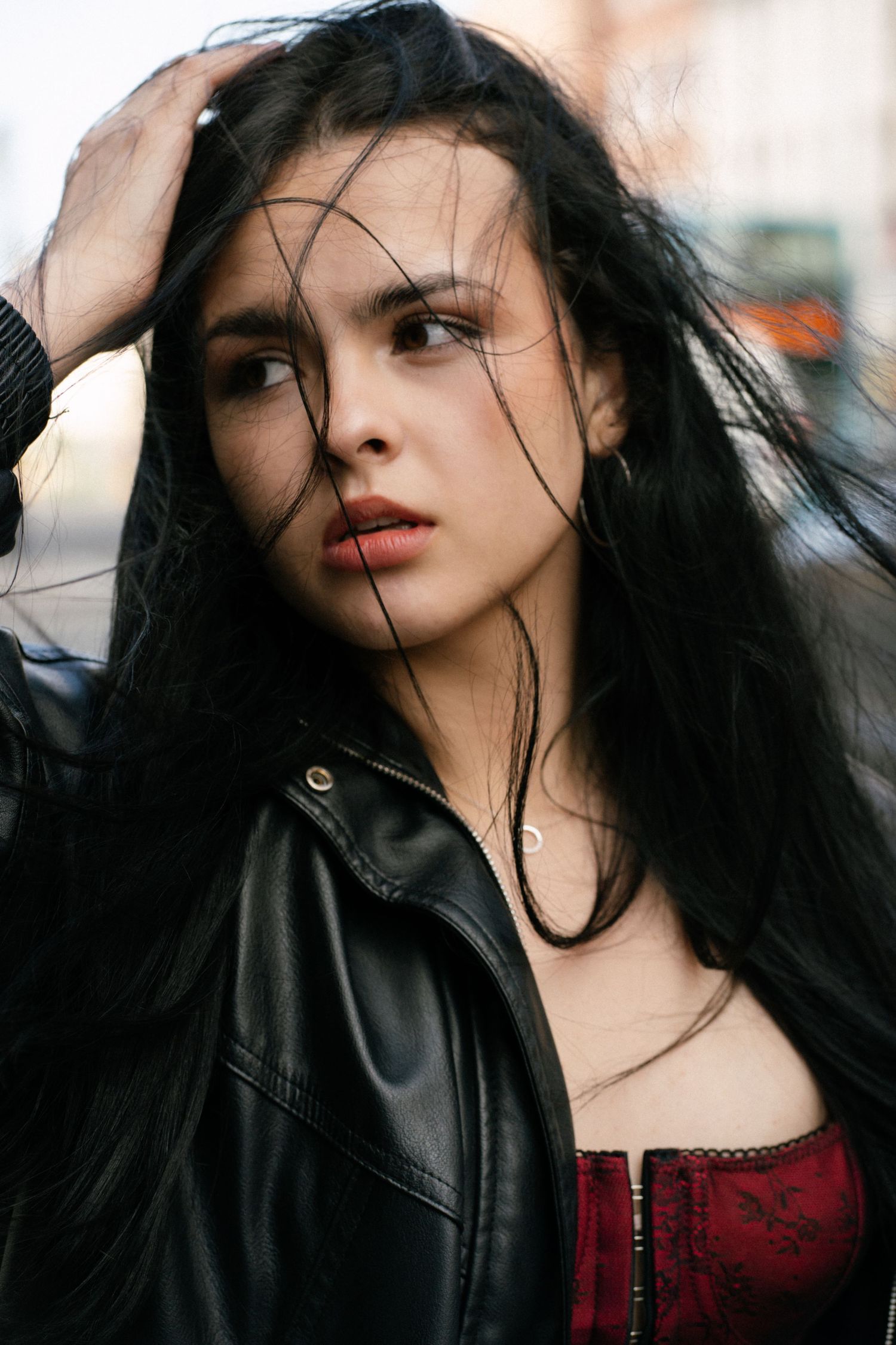 Young woman with long black hair in a red corset and black leather jacket poses outdoors in an urban setting.