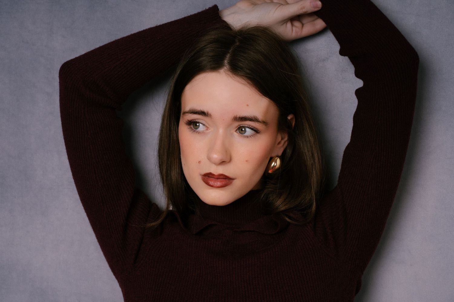 Brunette woman in dark turtleneck poses with arms raised against gray studio background.