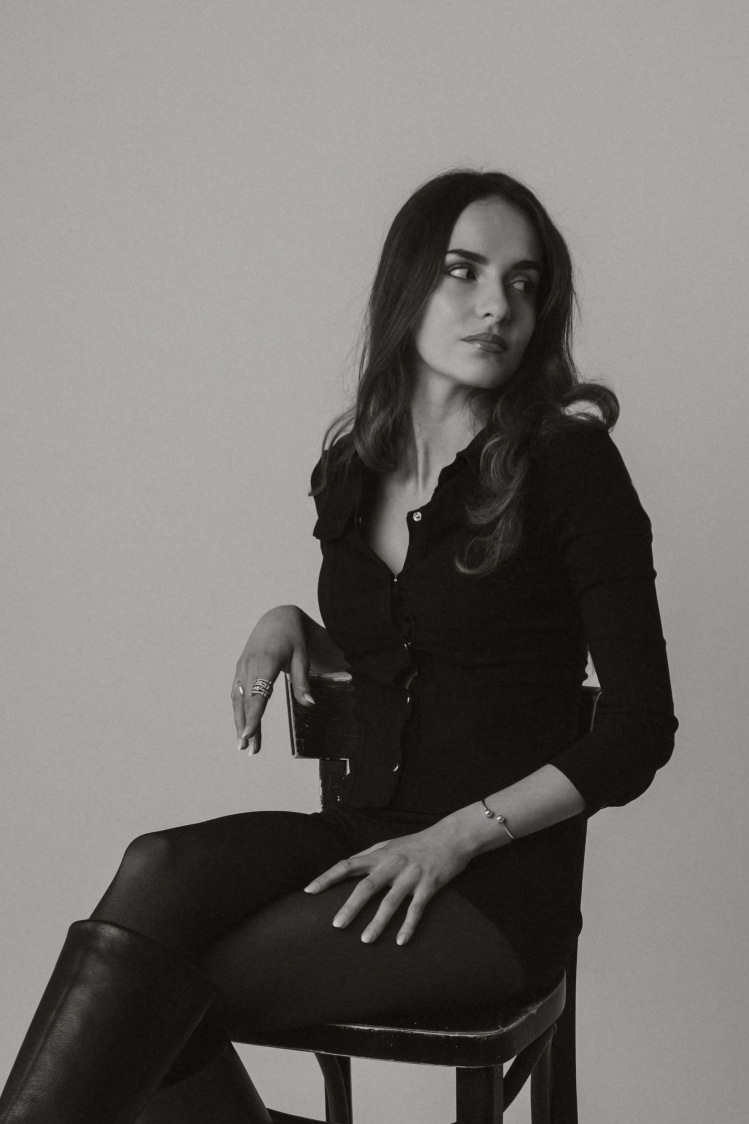 Black and white portrait of brunette woman in black shirt seated on chair wearing knee boots.