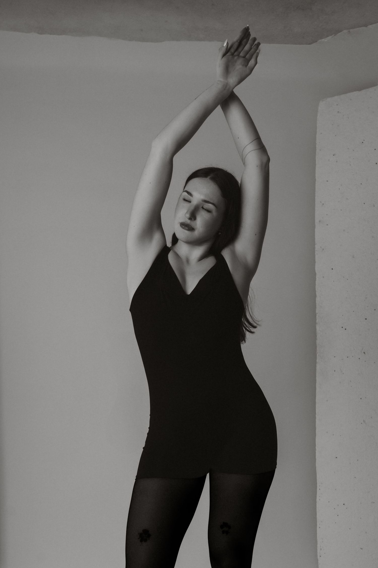 Black and white photo of woman in black bodysuit raising arms overhead, posing in studio corner.