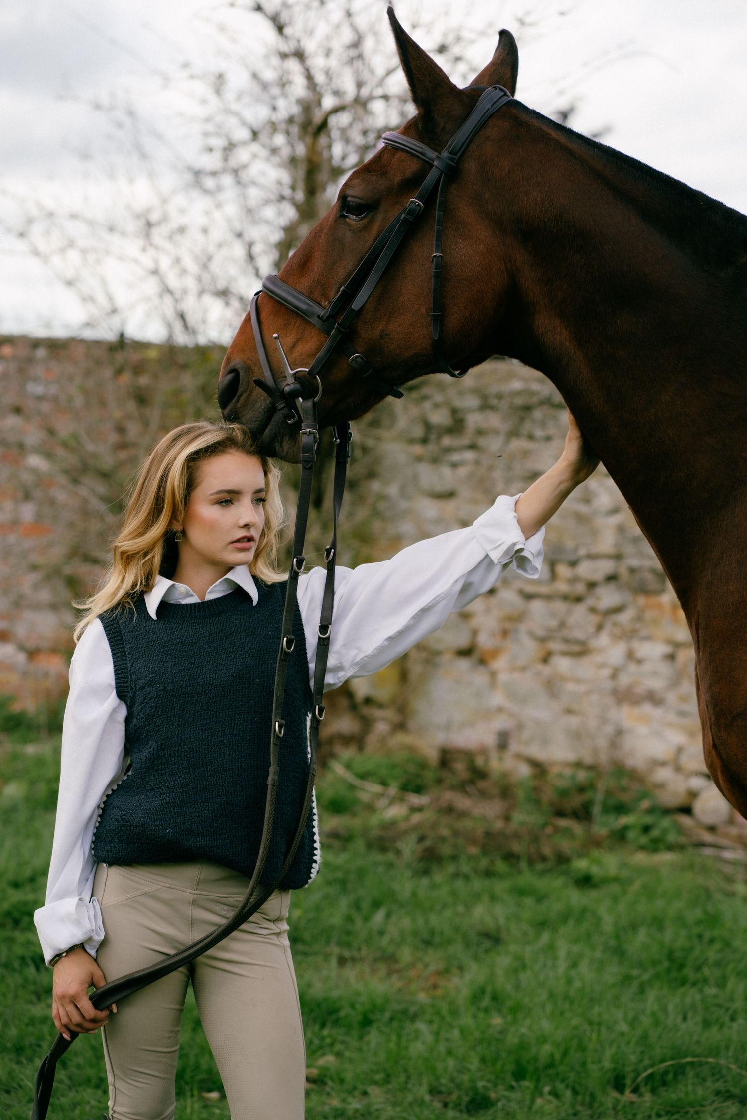 Young blonde woman in equestrian attire gently petting a brown horse outdoors near a stone wall.
