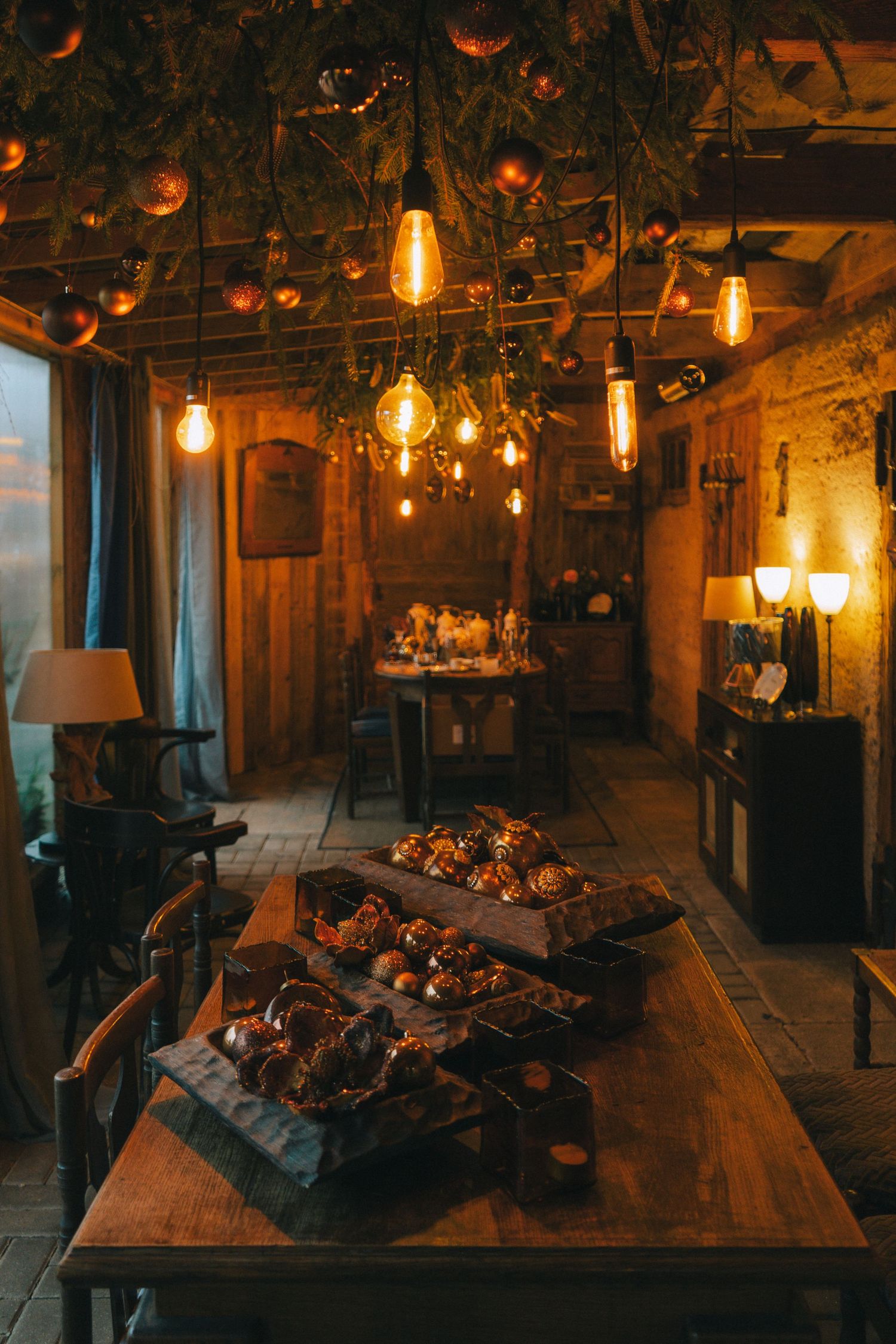 Rustic restaurant interior with Edison bulb lighting, wooden tables displaying decorative food arrangements, and lush greenery overhead.