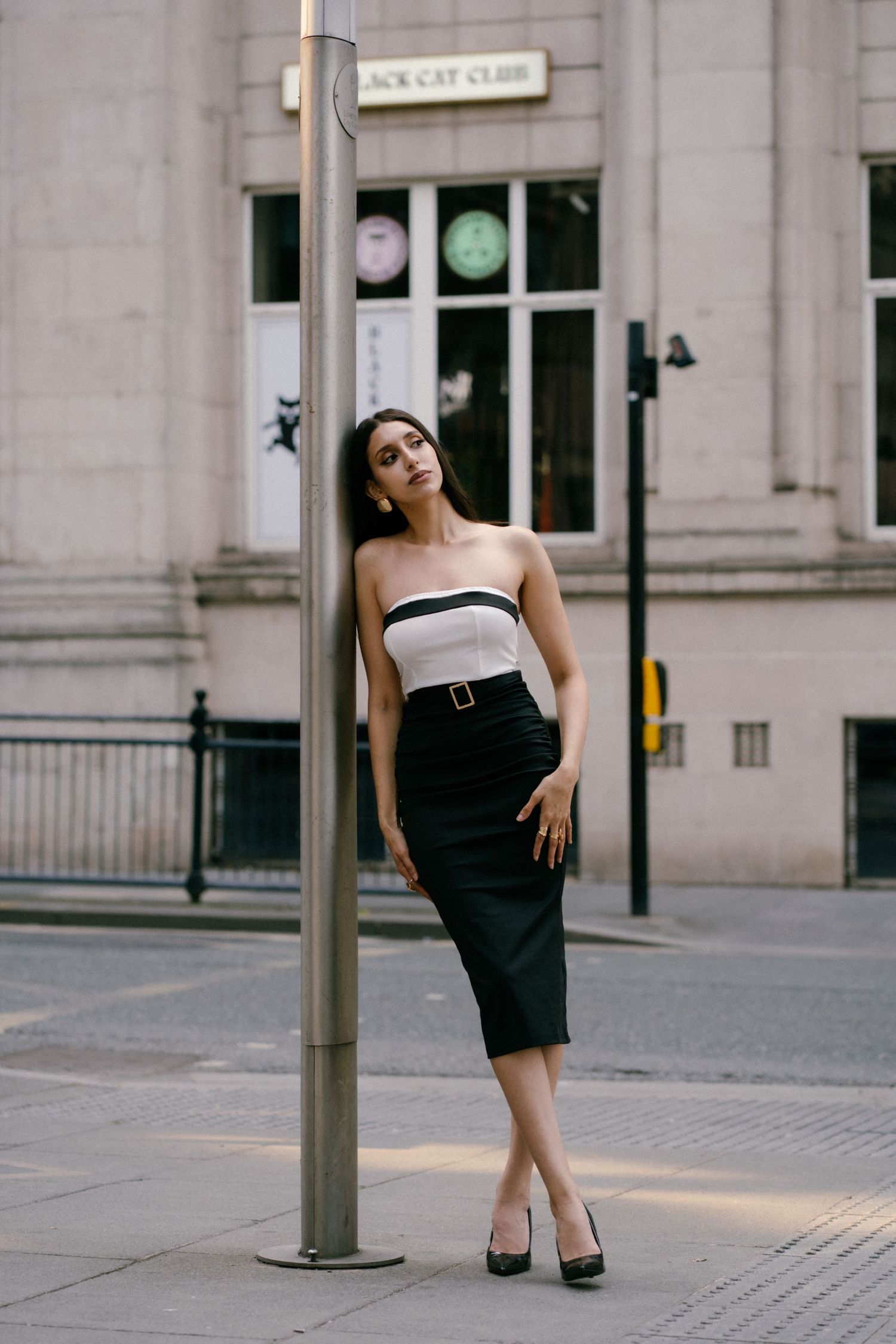 Woman in black pencil skirt and white strapless top poses elegantly by a street pole in an urban setting.