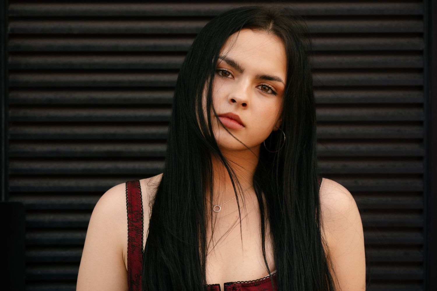 Young woman with long black hair and bold makeup poses confidently against a dark metal shutter background.