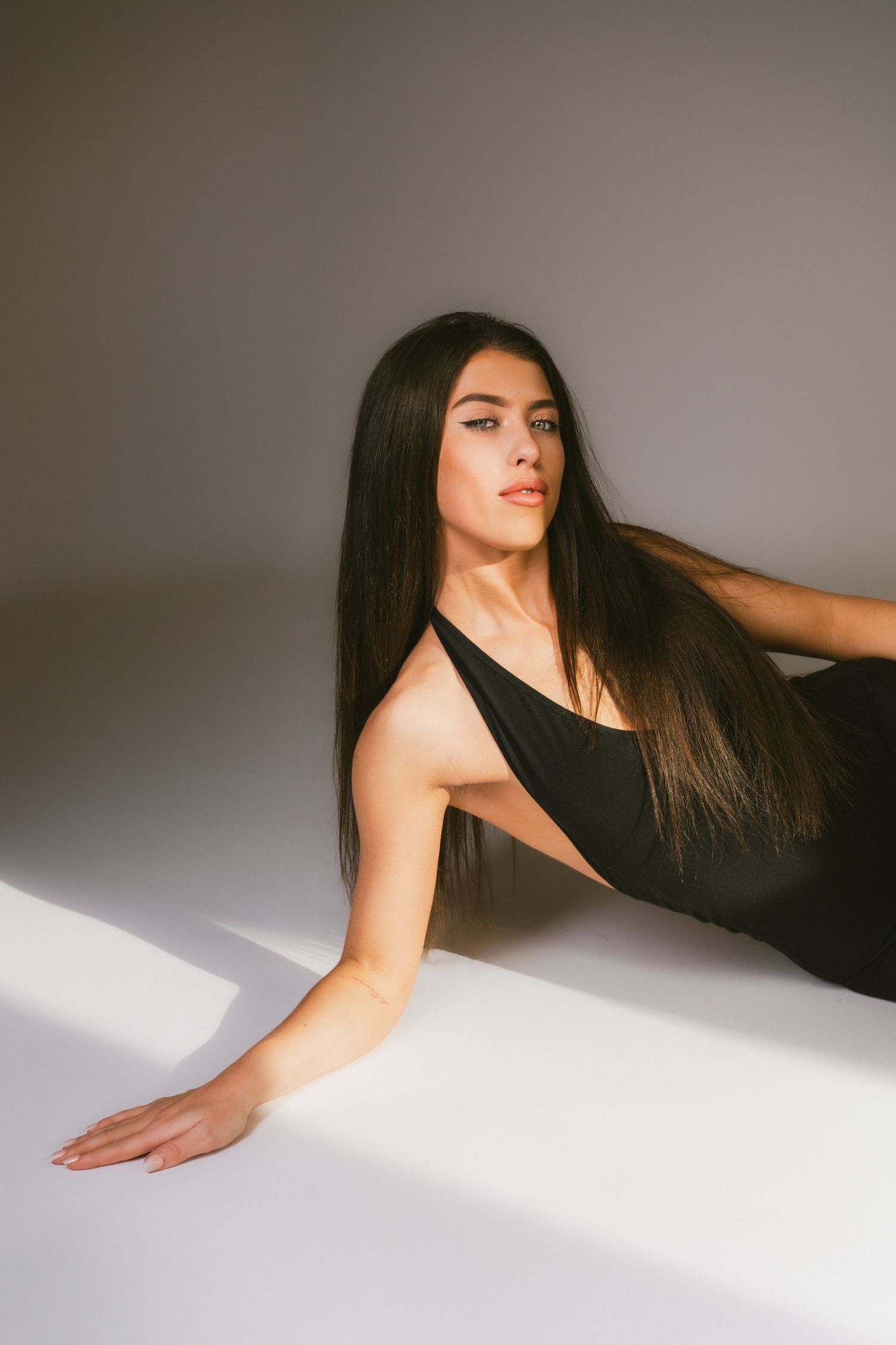 Woman with long dark hair in black asymmetric bodysuit reclining on floor in dramatic studio lighting.