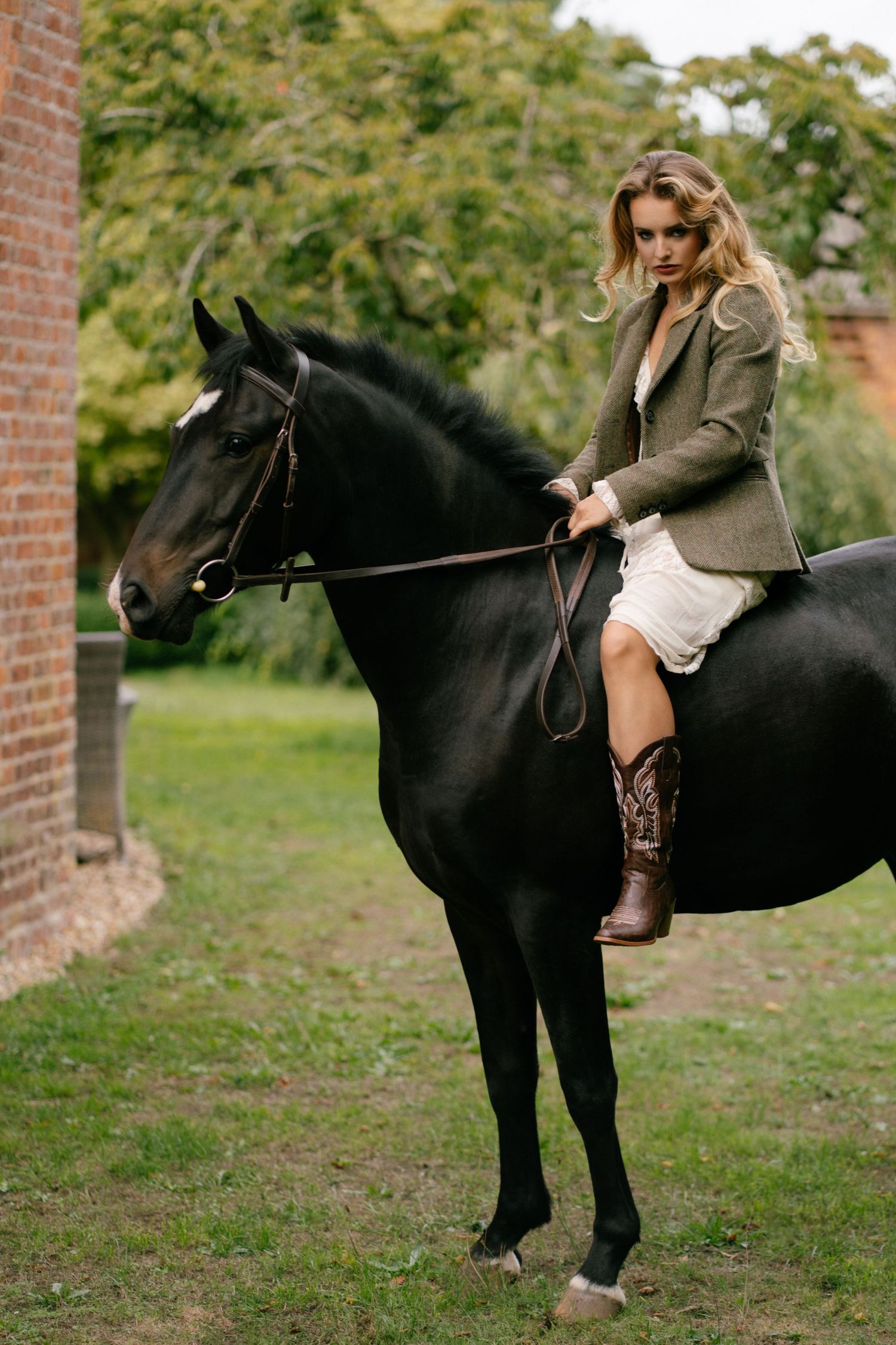A blonde woman in a blazer and cowboy boots rides a black horse near a brick building surrounded by green trees.