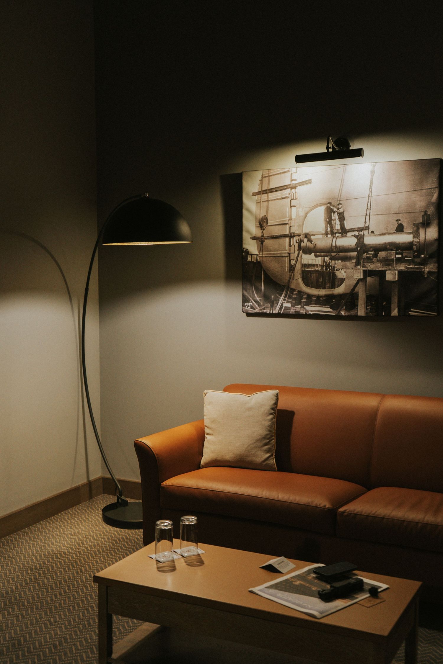 Modern hotel lounge with brown leather sofa, arc floor lamp, coffee table, and industrial artwork on gray wall.