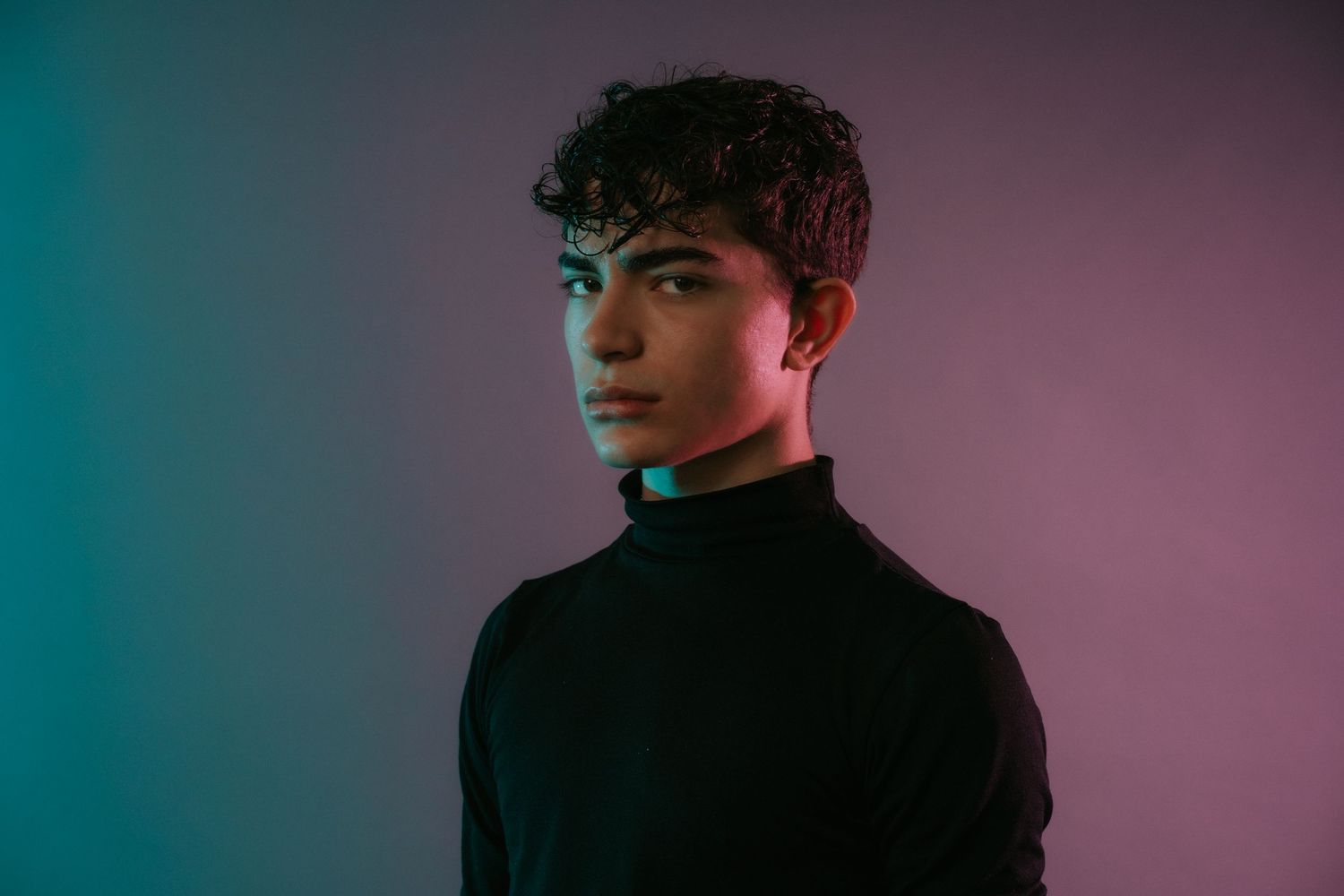 Young male model in black turtleneck under teal and pink studio lighting.