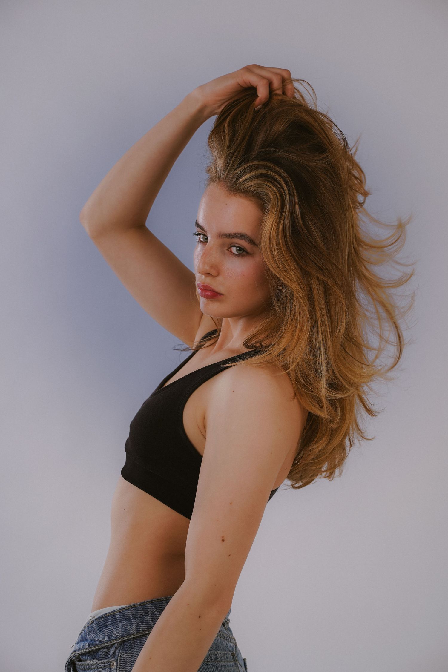 Young blonde woman in black crop top and jeans holding hair up against light gray background.
