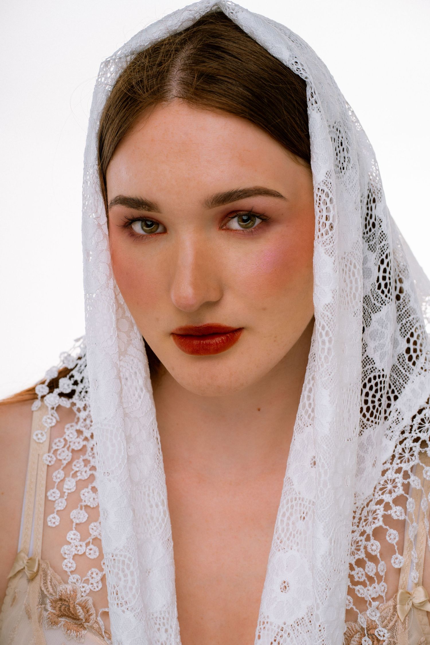 Elegant young woman with red lips wearing a delicate white lace veil over her auburn hair.