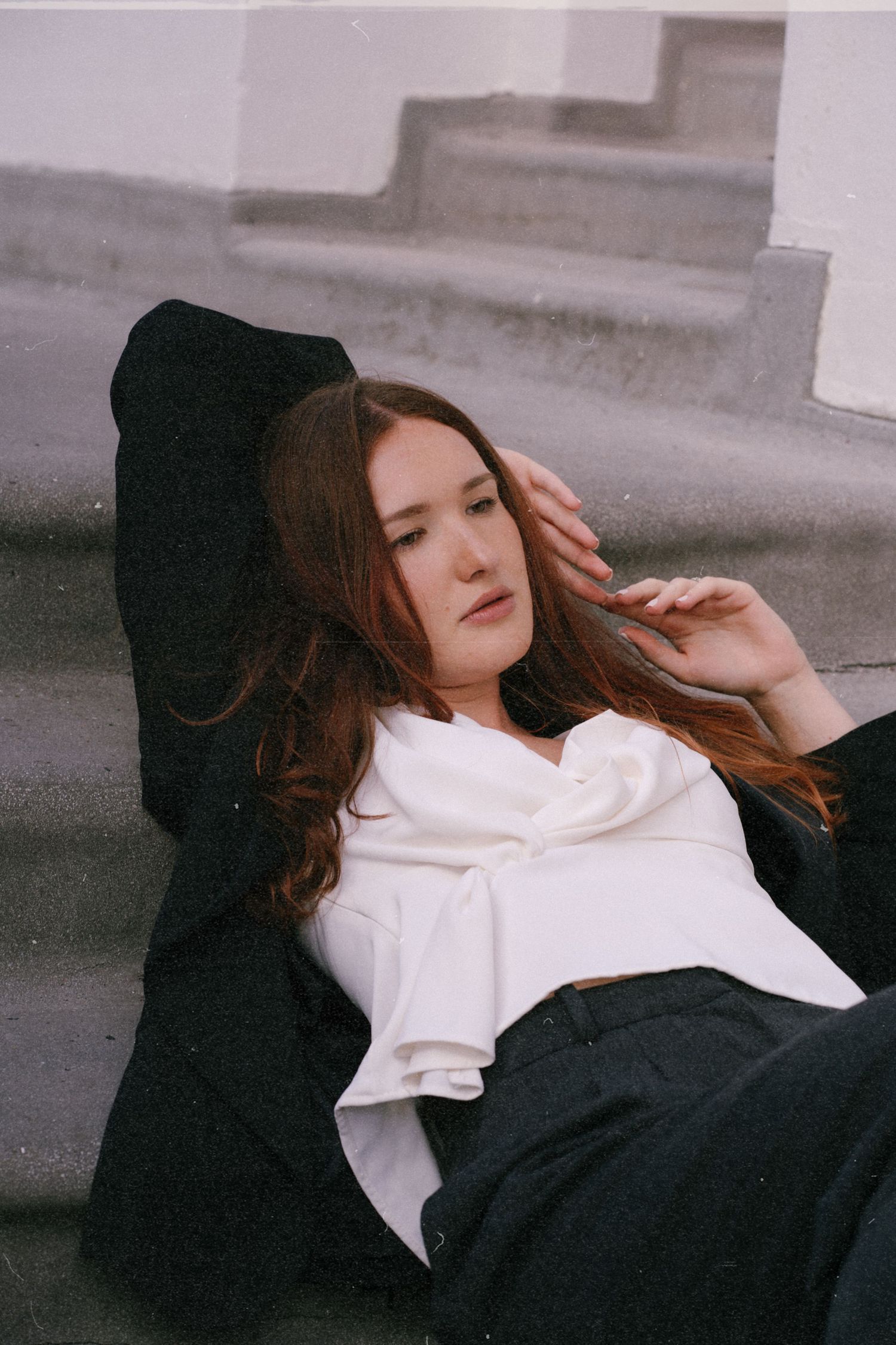 Young woman with auburn hair reclining on concrete steps wearing black outfit with white scarf.