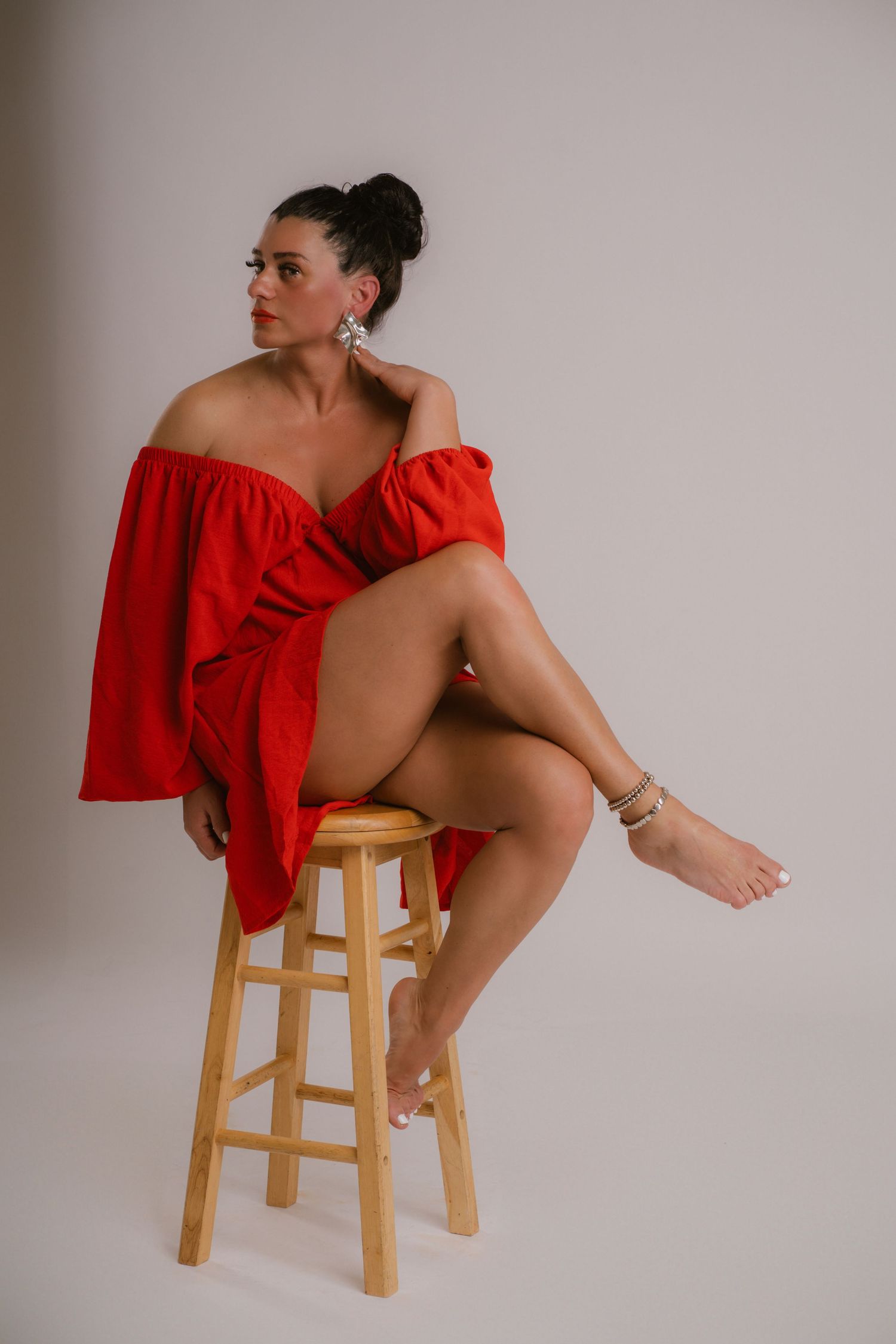 Woman in red off-shoulder dress sitting gracefully on wooden stool in studio portrait.