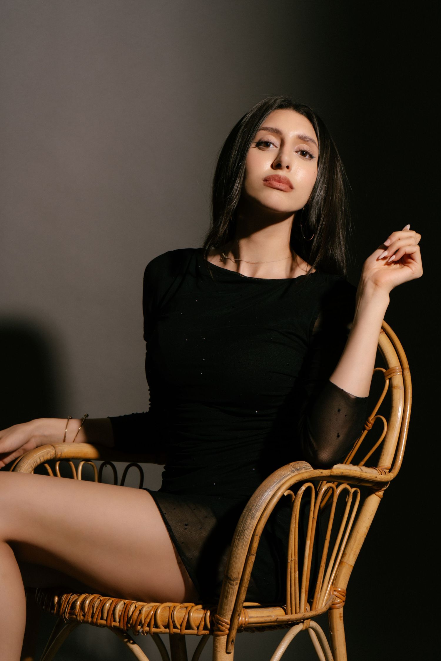 Dark-haired woman in black bodysuit seated gracefully in wicker chair, studio portrait.