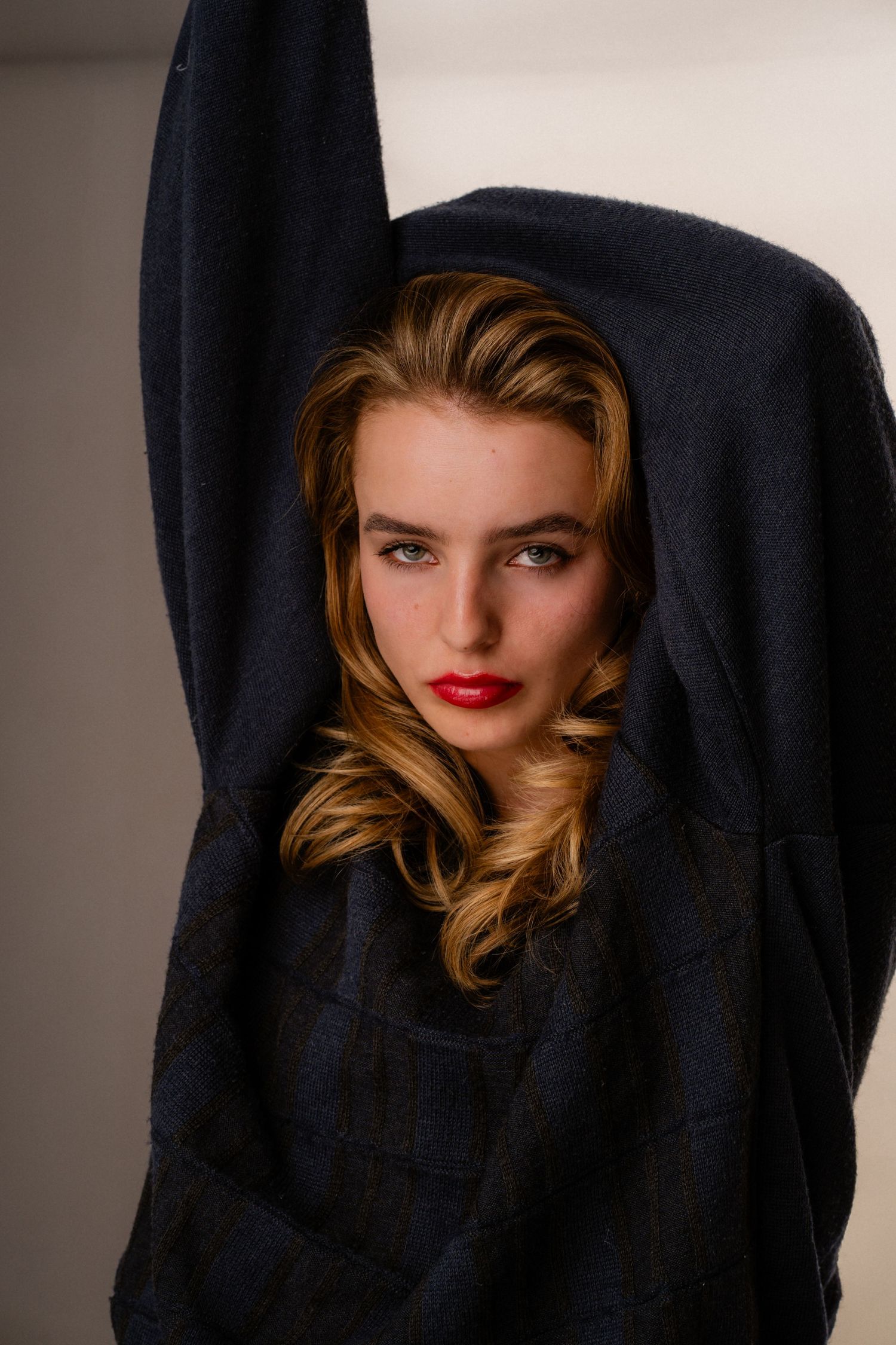 Young woman draped in dark blue fabric with arms raised, showcasing wavy blonde hair and red lips.