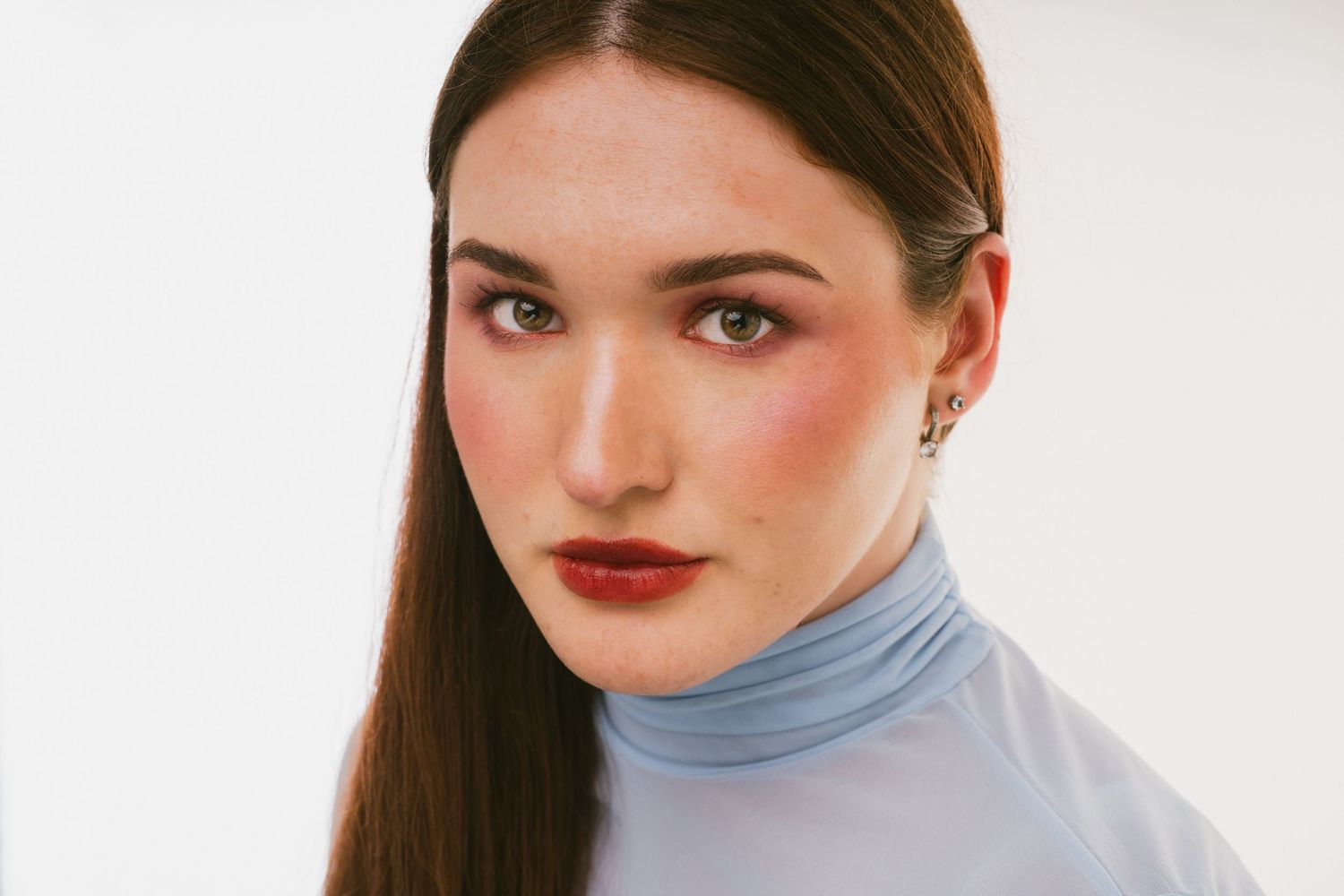 Close-up portrait of young woman with bold red lips and rosy cheeks in a blue turtleneck top.