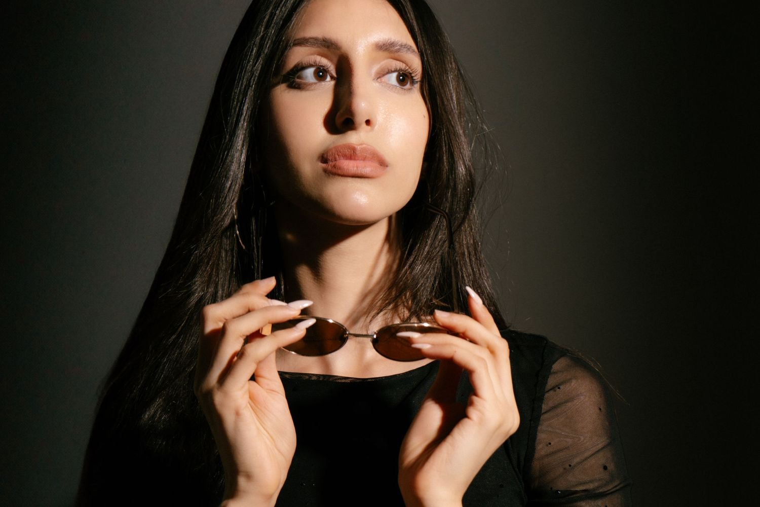 Dark-haired woman in black outfit holds sunglasses in dramatic low-key studio portrait.