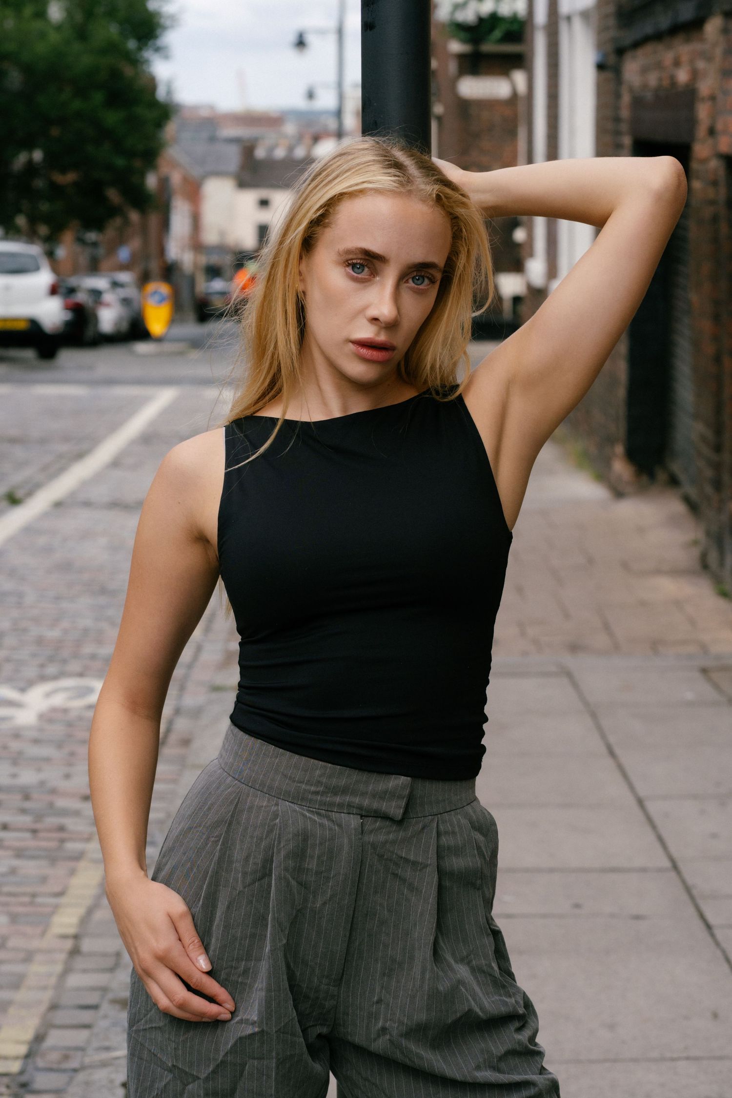 Young blonde woman in black tank top and gray pinstripe trousers posing confidently on a cobblestone city street.