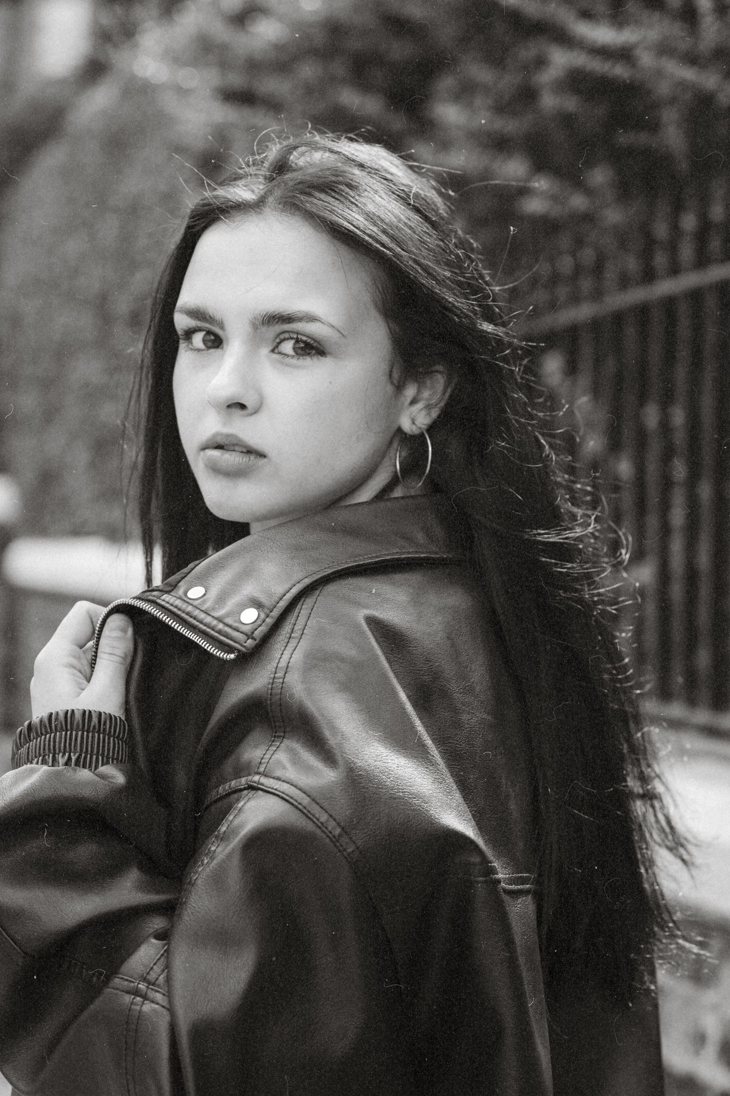 Black and white portrait of a young woman in a leather jacket with hoop earrings, posing outdoors near a fence.