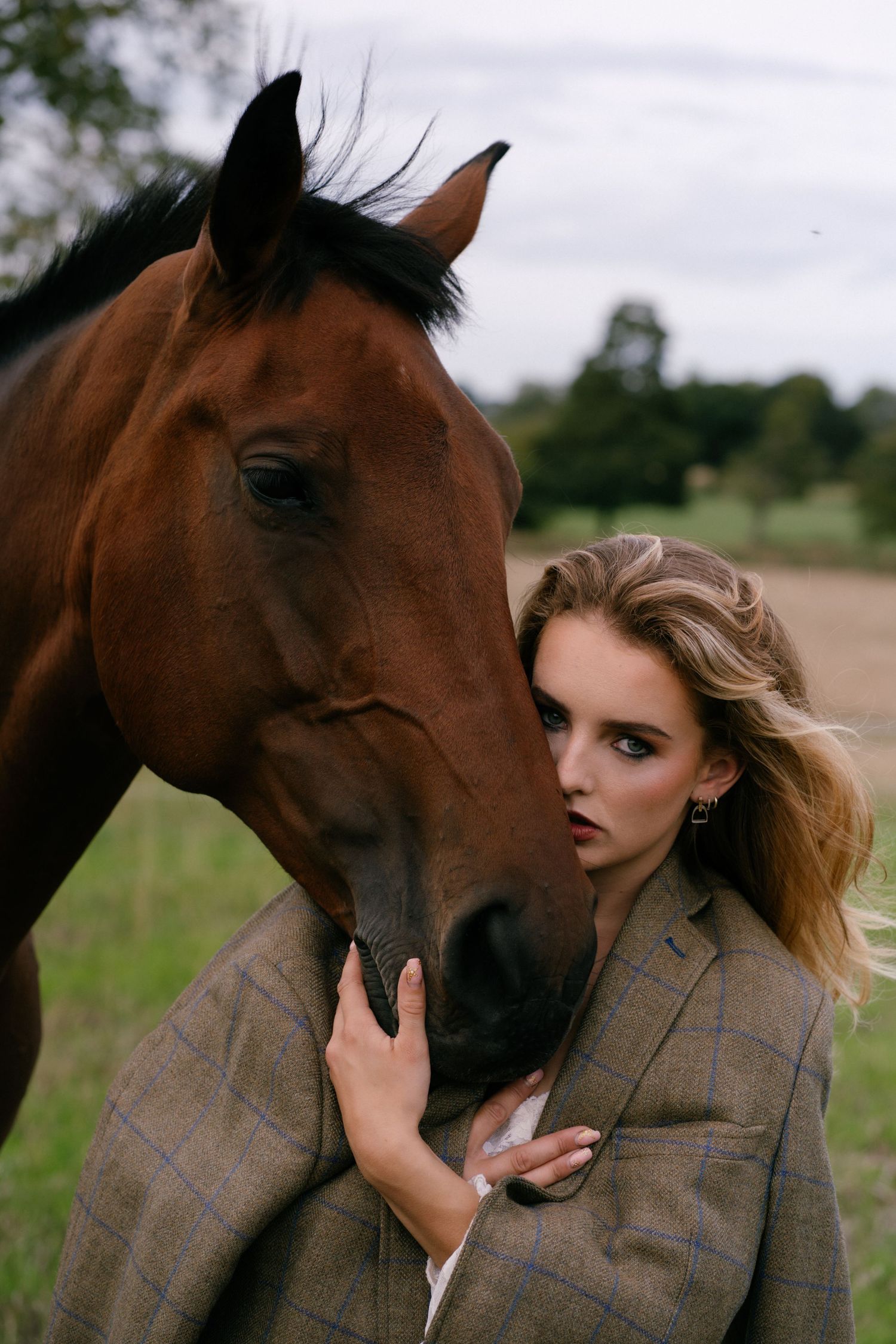 Blonde woman in plaid blazer tenderly embracing a brown horse in a field.