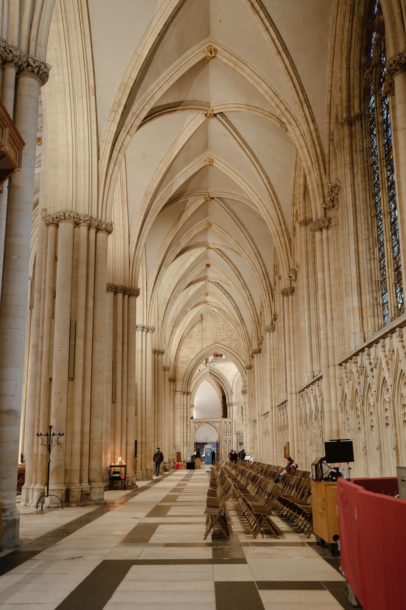 Gothic cathedral nave with soaring ribbed vaulted ceilings, stone columns, checkered floor, and rows of wooden pews.