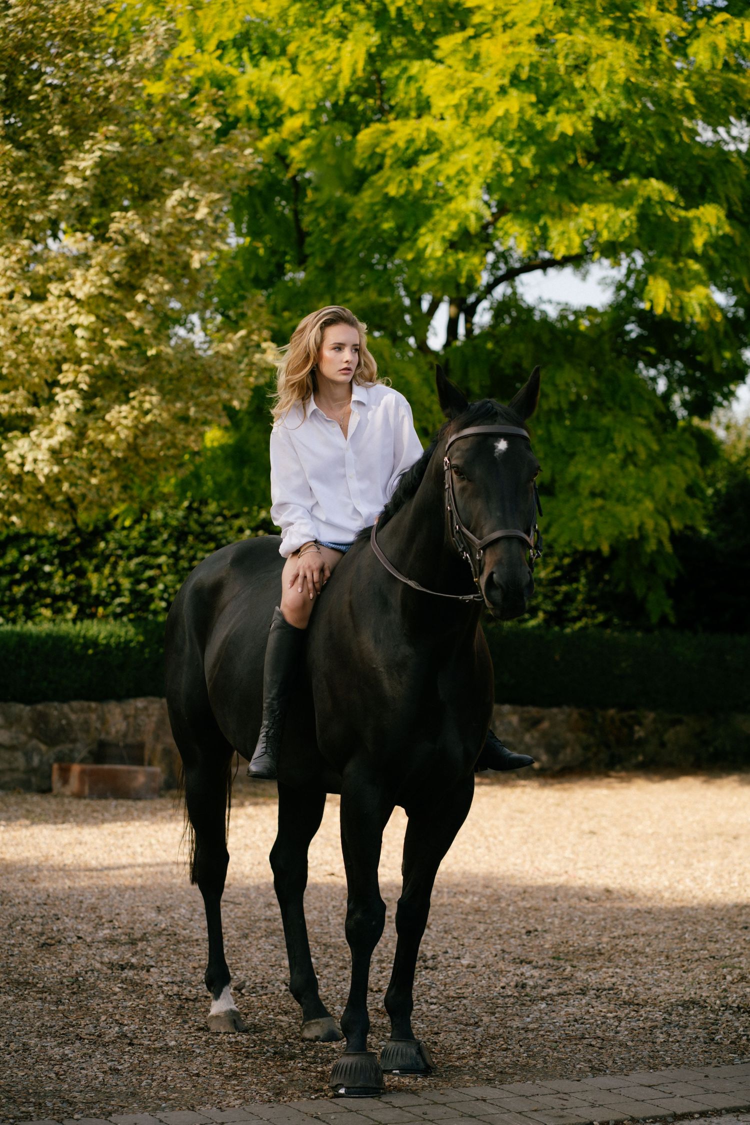 A blonde woman in a white shirt rides a dark black horse on a gravel path surrounded by lush green trees.