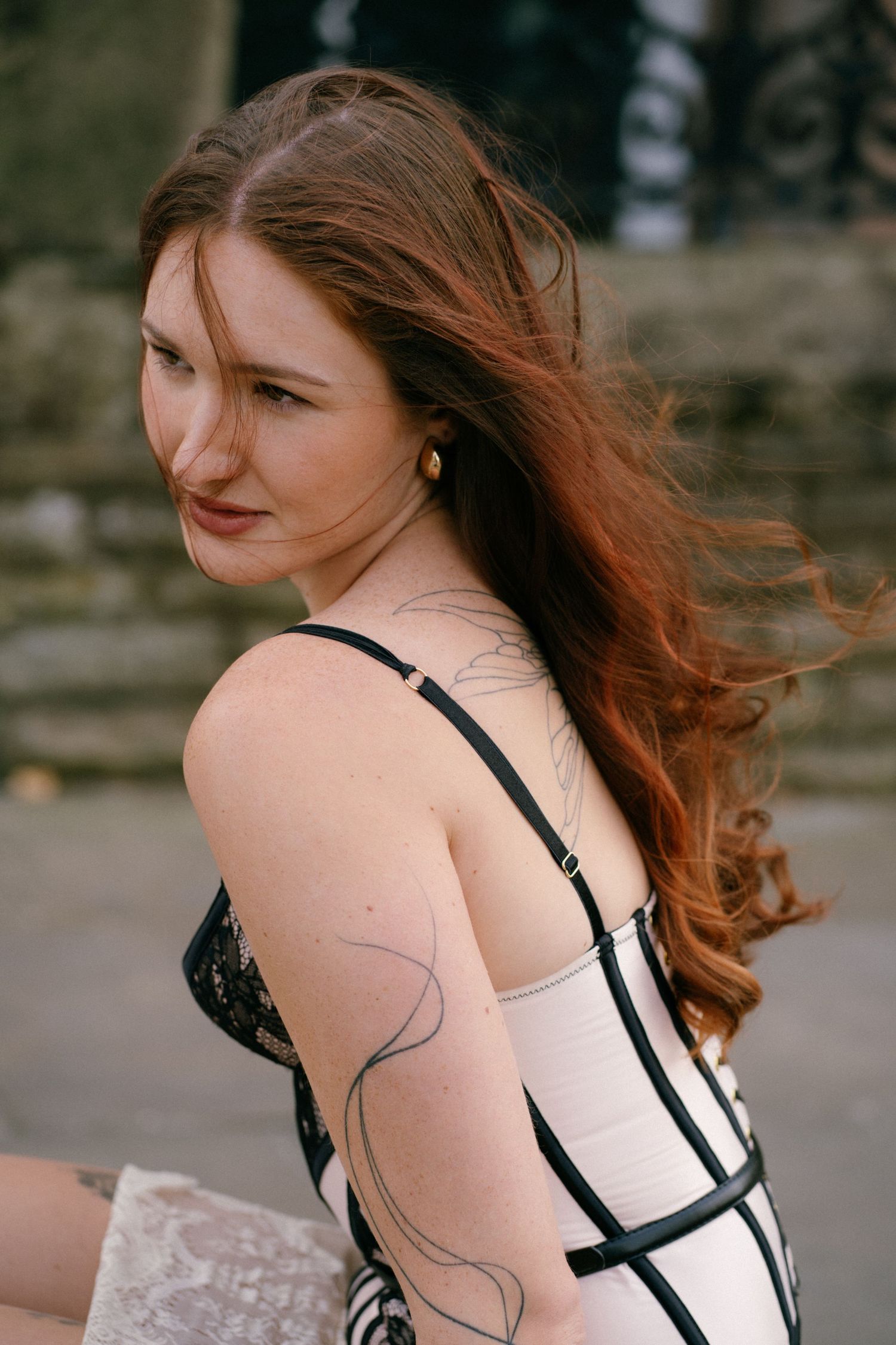 Young woman with long auburn hair posing outdoors in a black and white outfit with tattoos on her arm.
