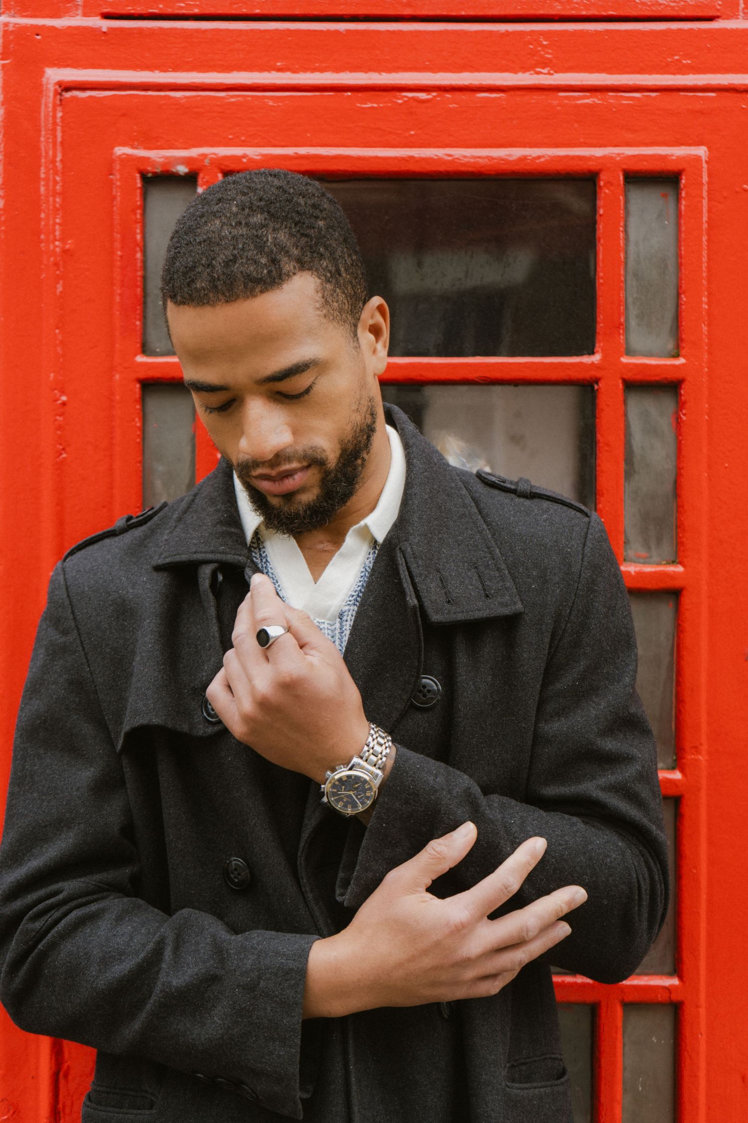 Stylish Black man in dark coat adjusting collar near iconic red London telephone box.